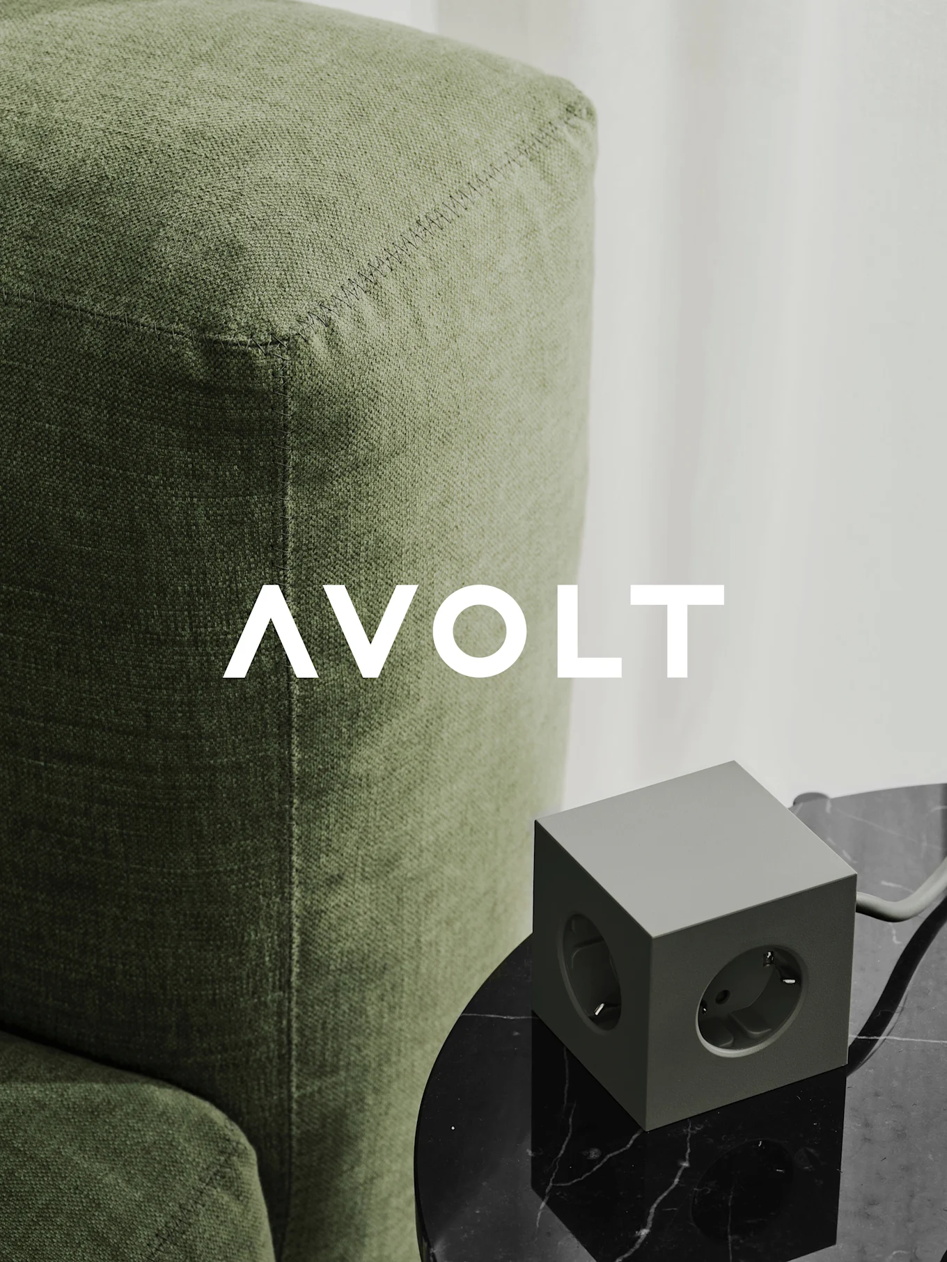 En grøn terningformet stikdåse med tre udtag på et sort marmorbord ved siden af en grøn sofa. AVOLT-logo.
