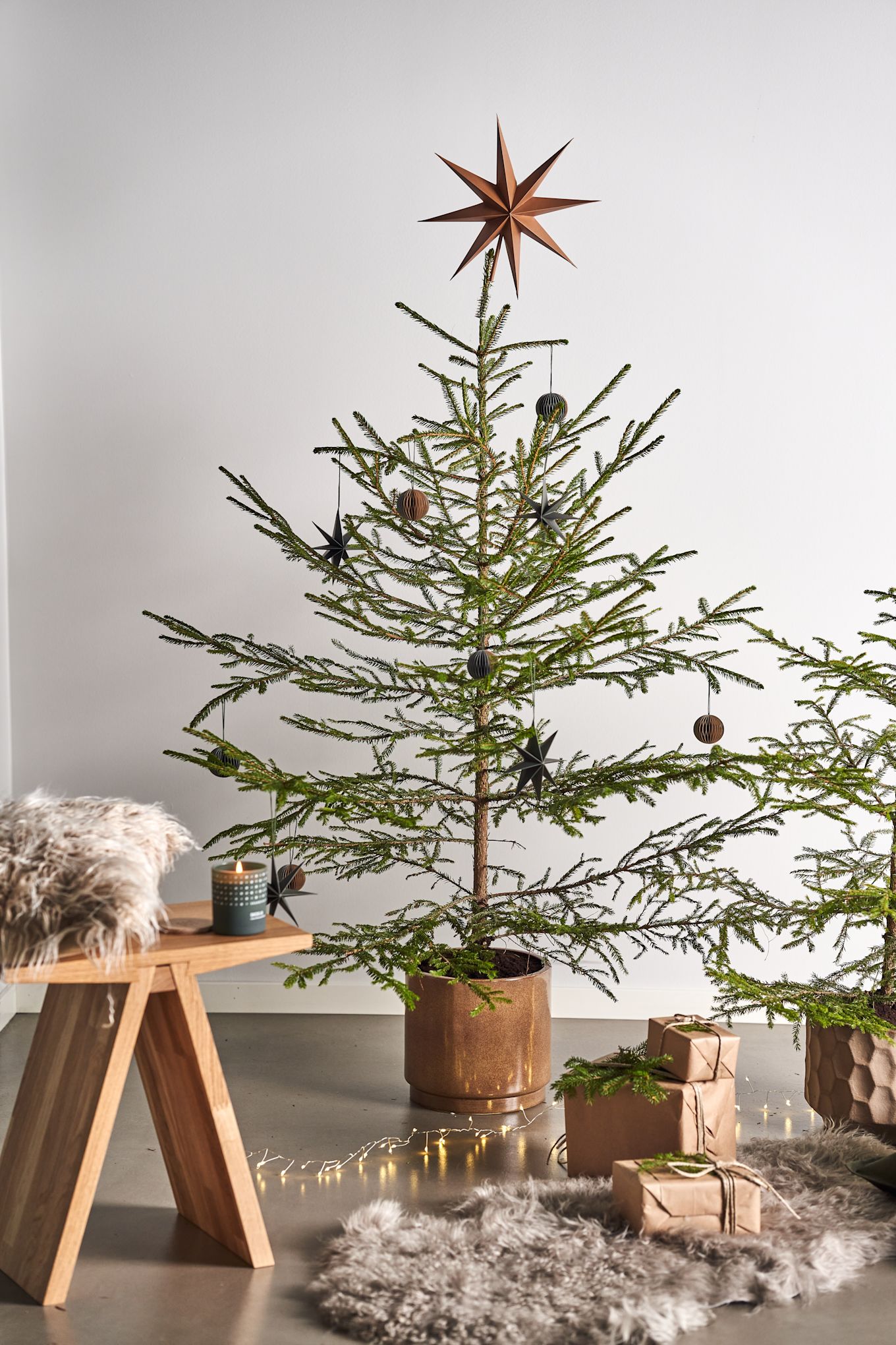 Pynt juletræet med juletræspynt til 2021 i 4 forskellige stilarter i henhold til Nest Trends - Nurture, Share, Boost og Cultivate. Her ses Angle skammel fra Muubs med A Skandinavisk duftlys i Forest duften ved siden af pakker på et fåreskindstæppe. 
