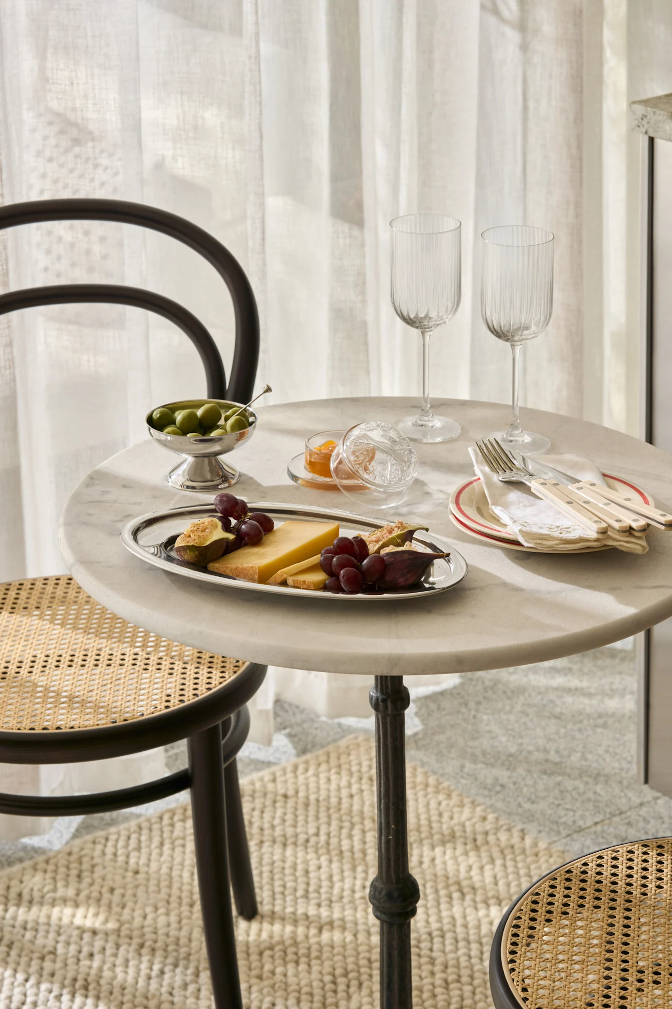 Et marmorbord dækket med ost, figner, druer, oliven, syltetøj, glas og bestik, med fletstole.