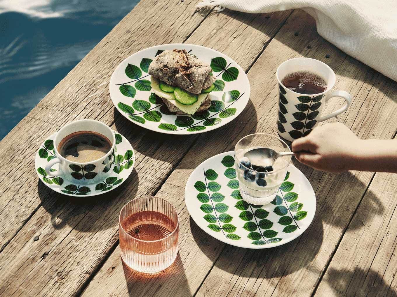 Morgenmad på træmole: Tallerkener med bladmønster, sandwich, kaffe, te, yoghurt med blåbær og lyserød drik.