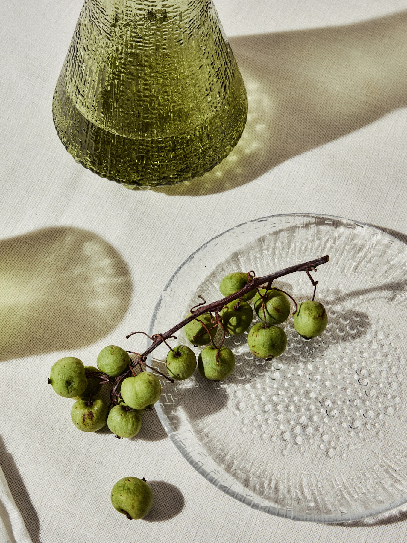 Grøn tekstureret glasvase og en klar tallerken med små grønne frugter på en vinranke.