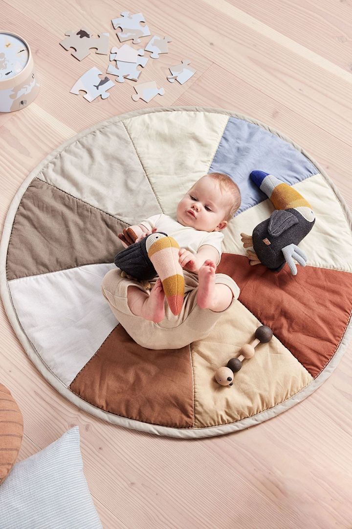 Trine Toucan bamsen fra OYOY ligger ved siden af en baby på Moni-børnetæppet. Bamsen er udelukkende lavet af genbrugsmaterialer, hvilket ikke kun gør den til en brugbar, men også en bæredygtig gave til babyshower.