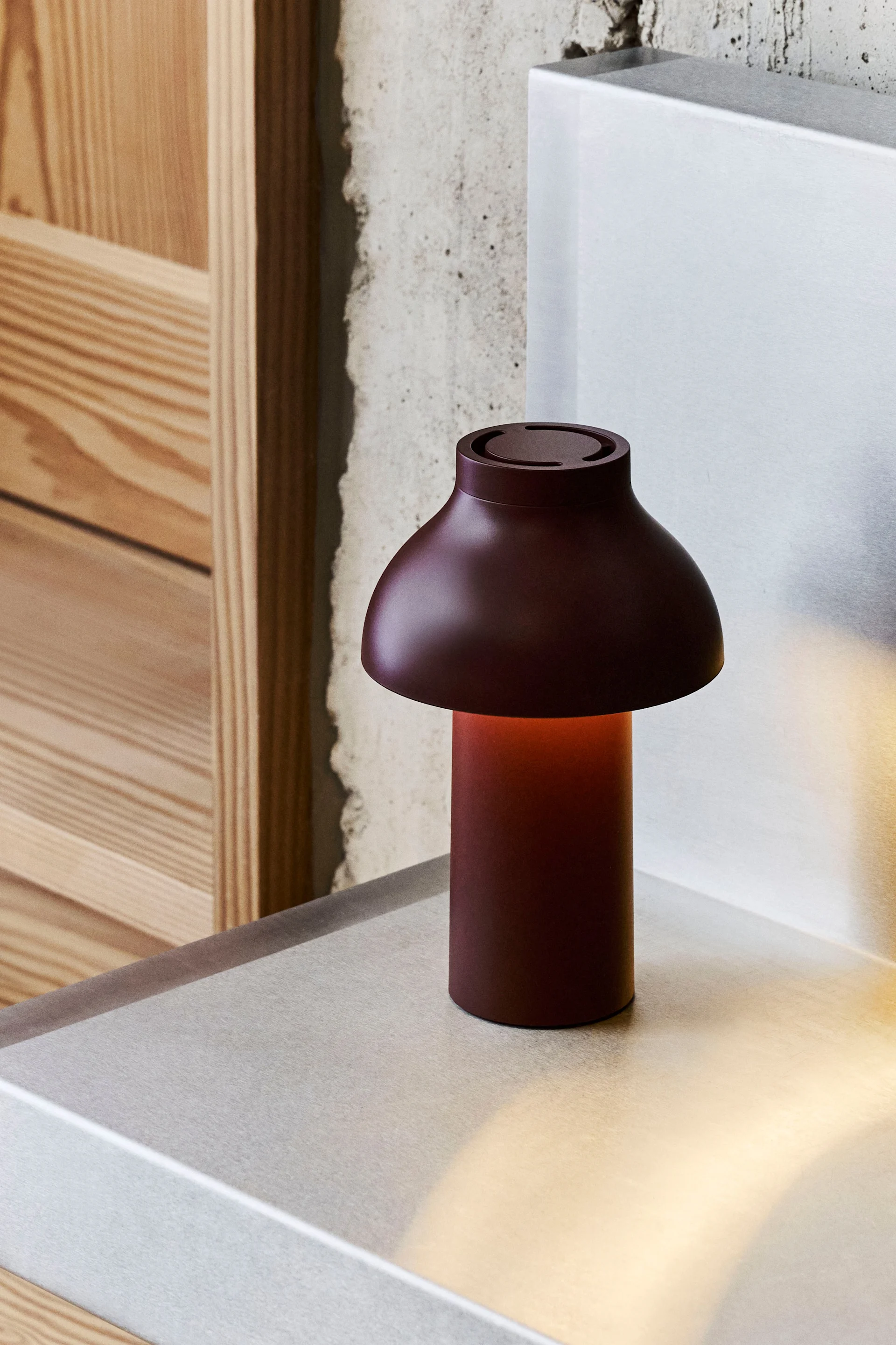 En mørkerød champignonformet bordlampe med varmt skær står på en metallisk overflade, med beton og træ i baggrunden.