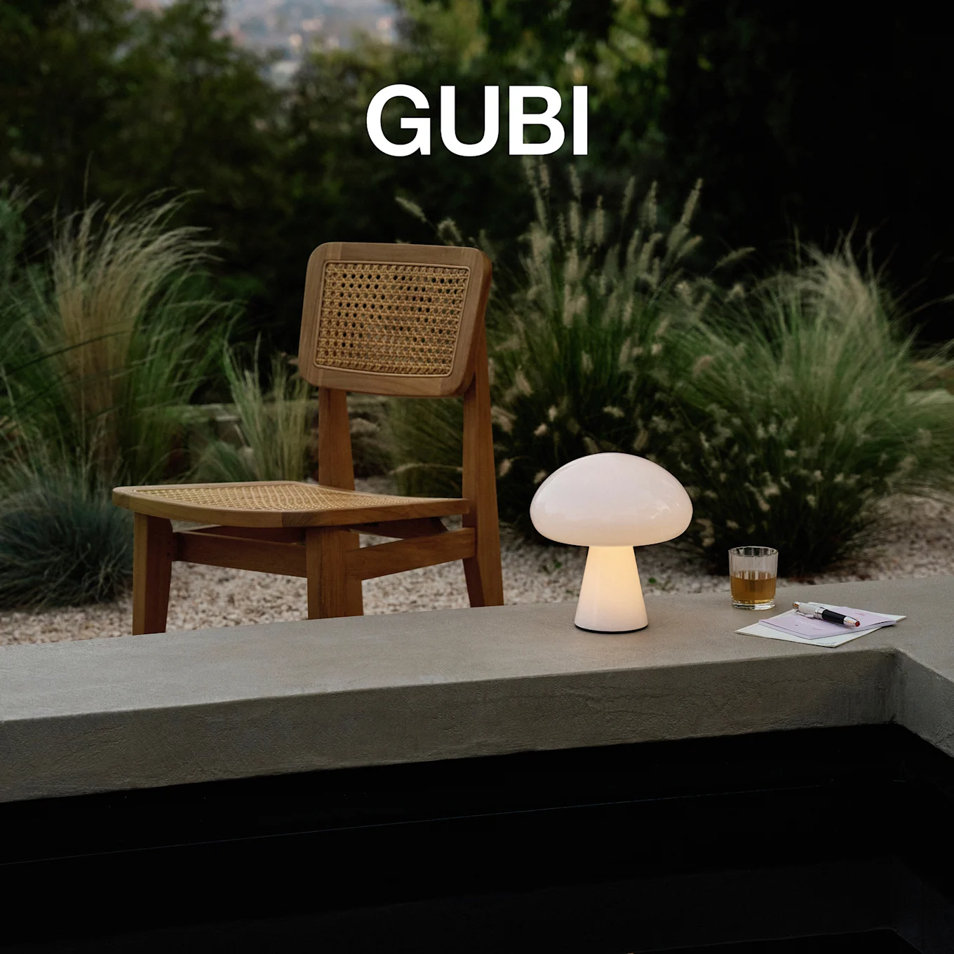 GUBI logo. En oplyst champignonformet bordlampe, et glas og en pen på en betonafsat, med en træstol og grønne planter.