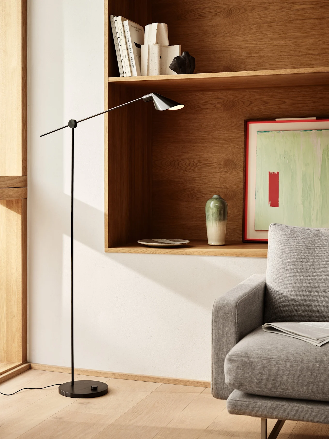 En moderne stue med en slank sort gulvlampe, træhylder med bøger og dekoration, og en del af en grå sofa.