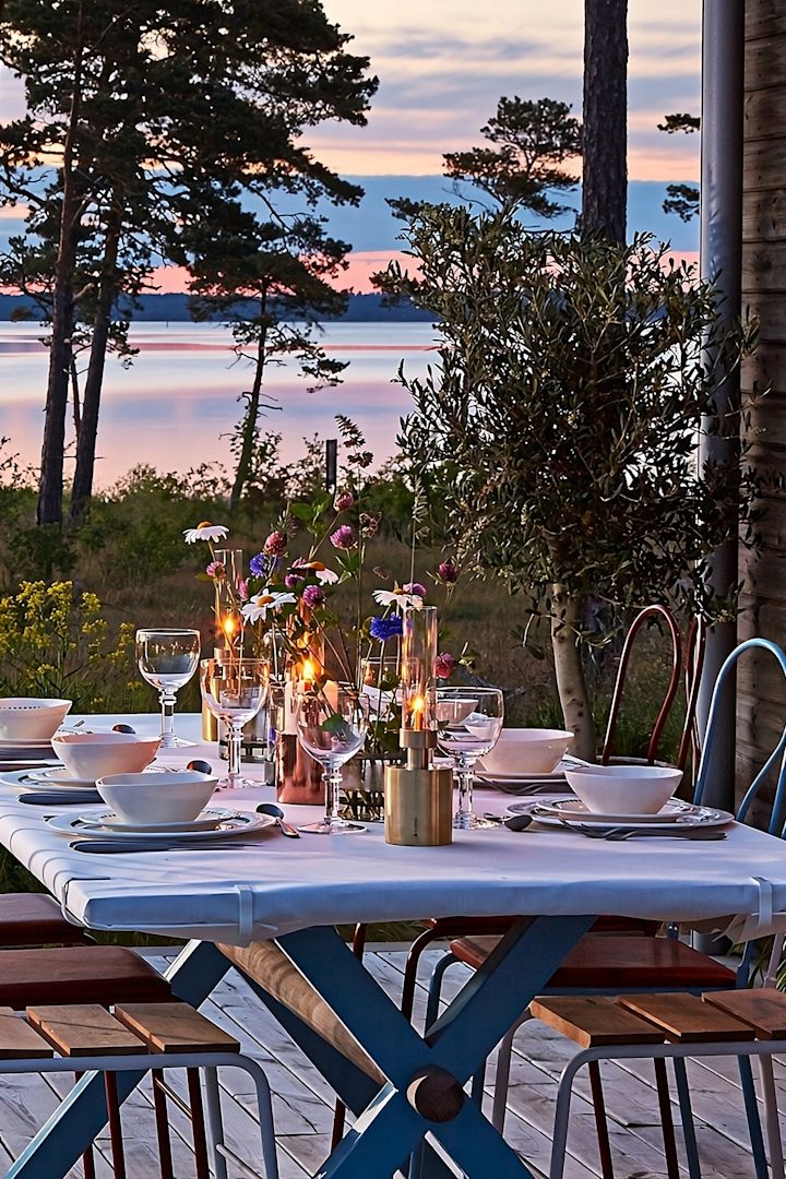 Lanterner og lygter til udendørs brug - her ses et træbord på en terrasse med havet i baggrunden i solnedgangen. Bordet er dækket og dekoreret med blandt andet Patina mini-olielampen fra Klong.