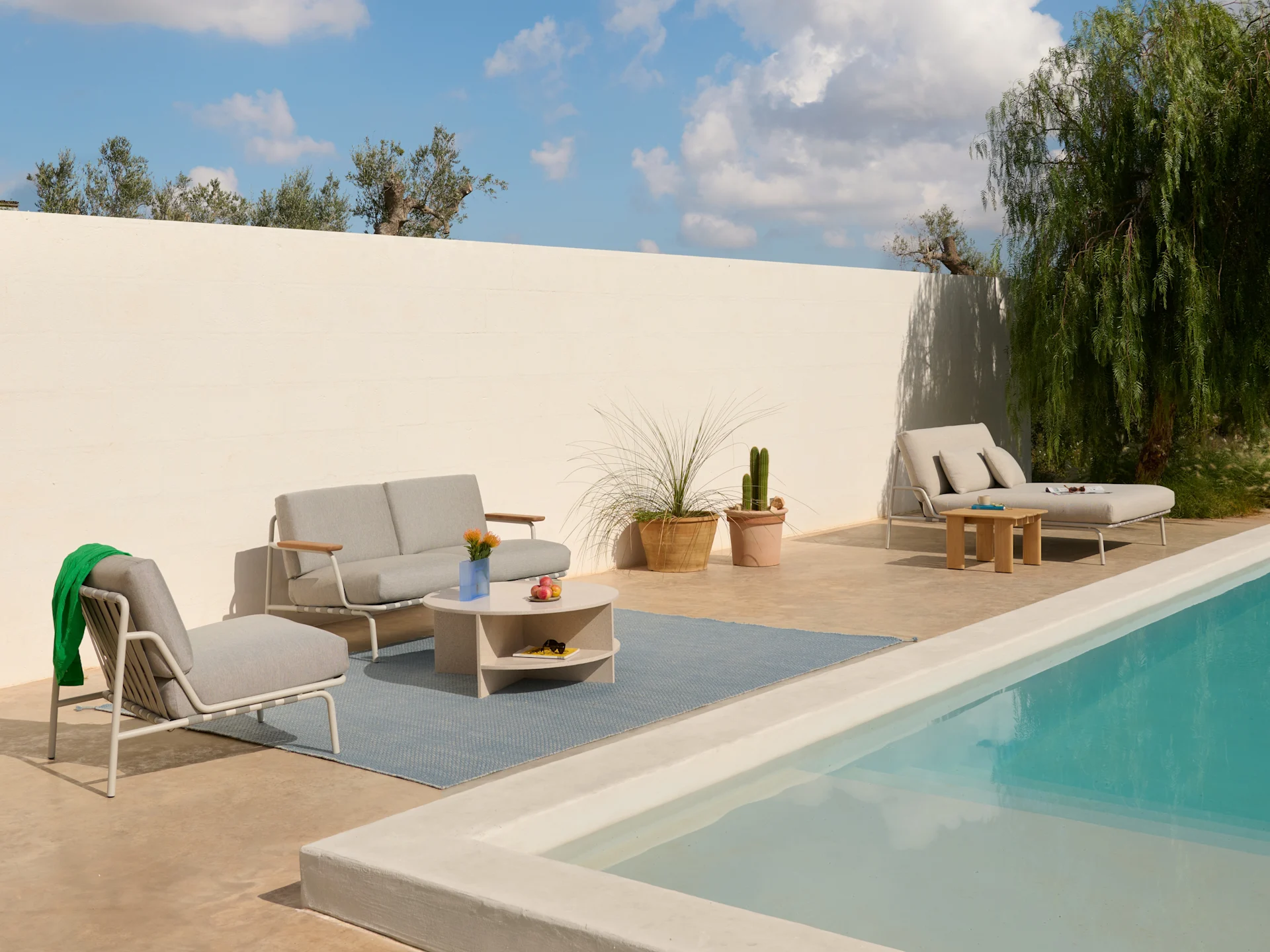 Udendørs terrasse med lysegrå sofa, lænestole, sofabord på blåt tæppe og en liggestol ved en pool.