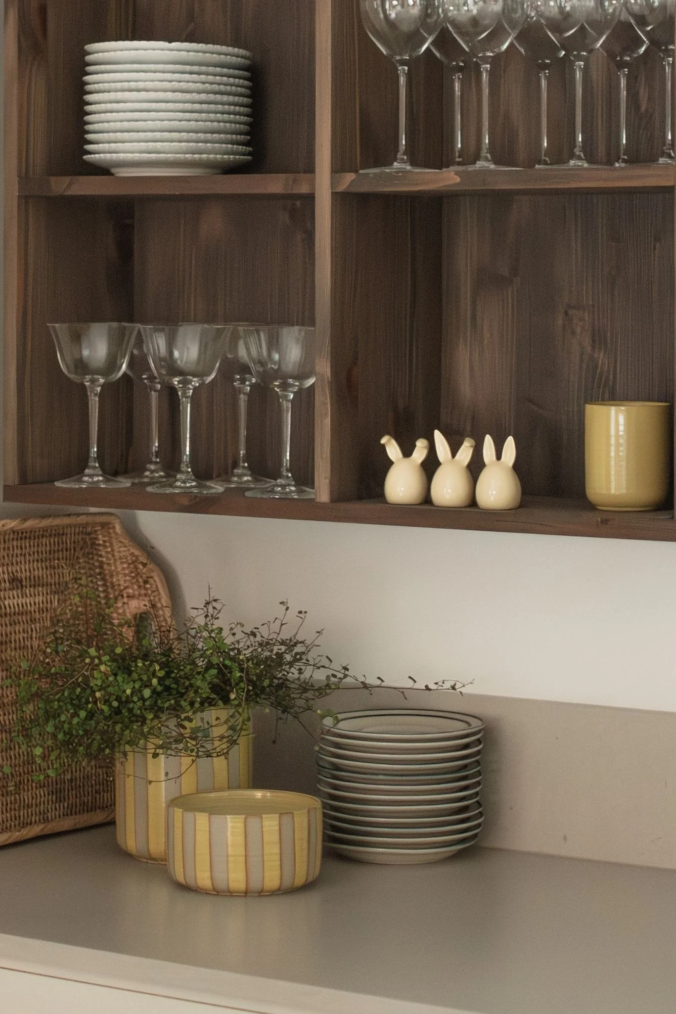 Træhylder med stablede hvide tallerkener, forskellige glas, tre keramiske kaniner og et gult krus. På disken en flettet kurv, planter i stribede potter og flere tallerkener.