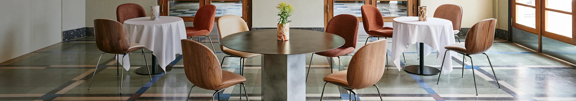 Stilfuldt caféinteriør med et stort centralt bord, to mindre borde med hvide duge og diverse stole på et mønstret gulv.