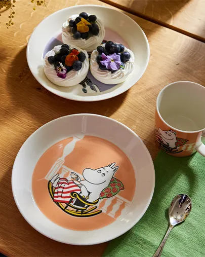 Moomintroll tallerken, krus og ske ved siden af en tallerken med marengs med blåbær og blomster på et træbord.