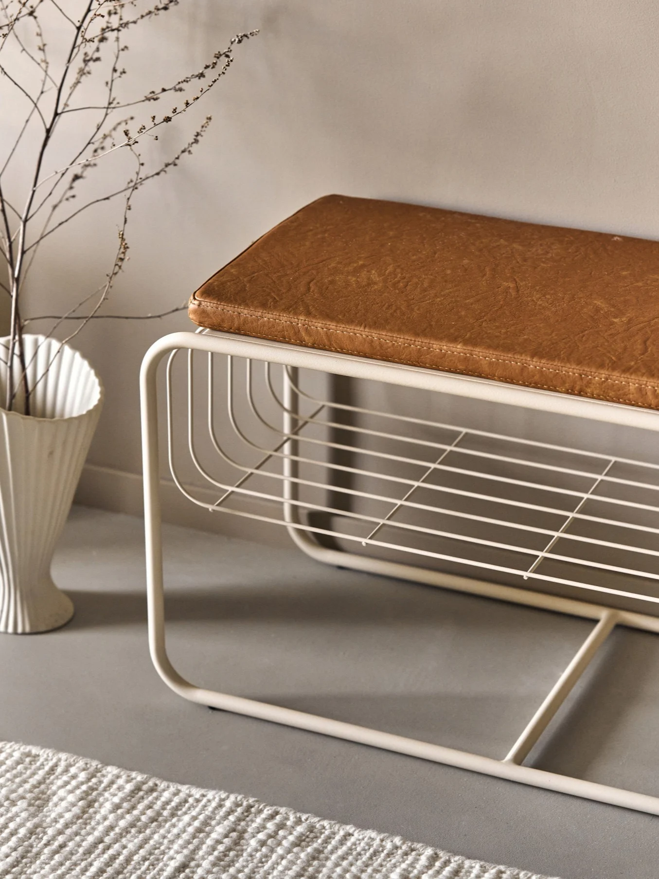 Moderne beige metalbænk med brun kunstlæderhynde, med en åben lamelhylde. En hvid vase med grene og et tæppe er i nærheden.