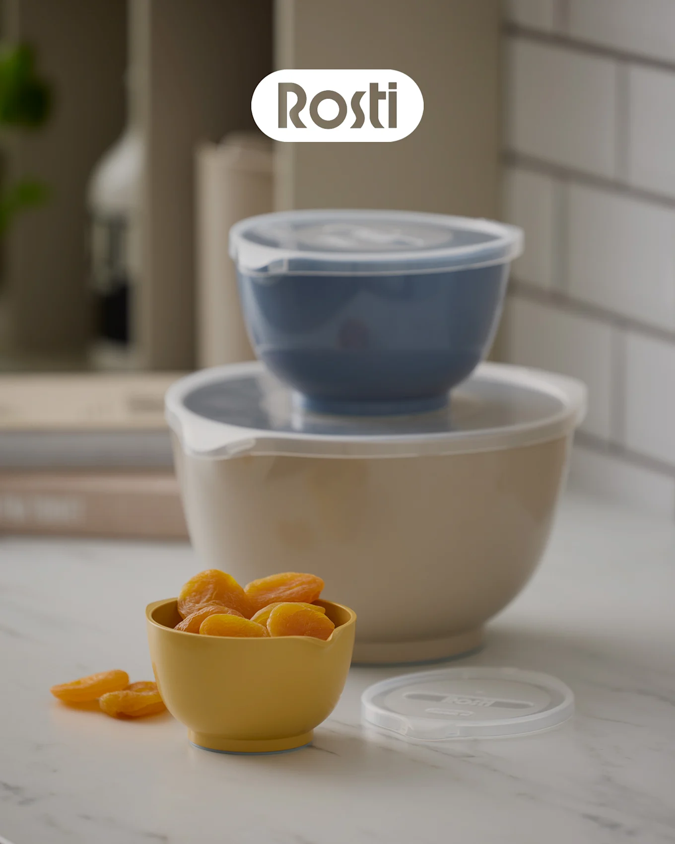 Tre Rosti skåle på en disk: en beige skål med en blå stablet ovenpå, begge med låg, og en lille gul skål fyldt med tørrede abrikoser.