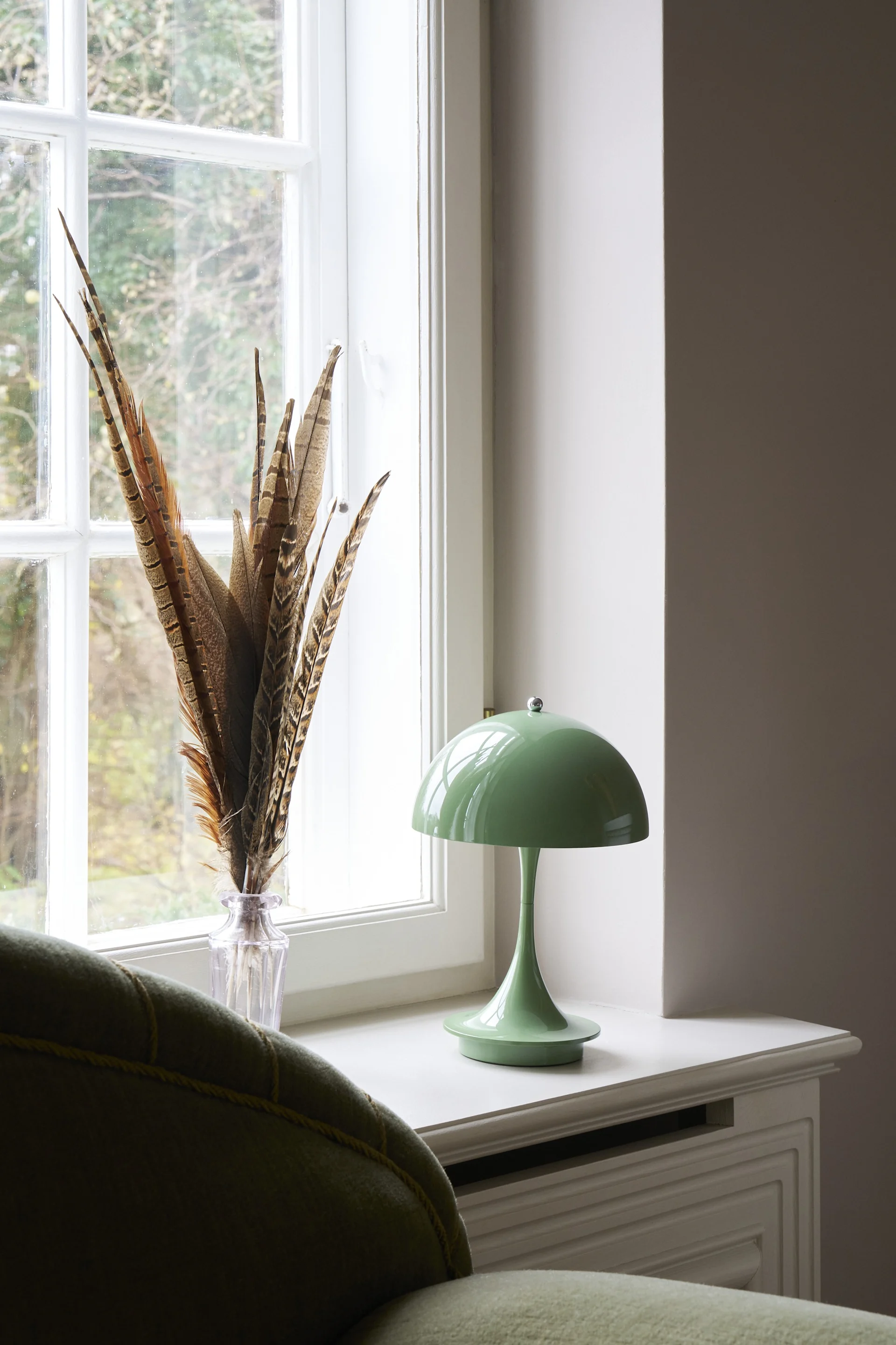 En grøn svampelampe og vase med fjer på en vindueskarm ved siden af en grøn sofa, med en kande af rattan i baggrunden.