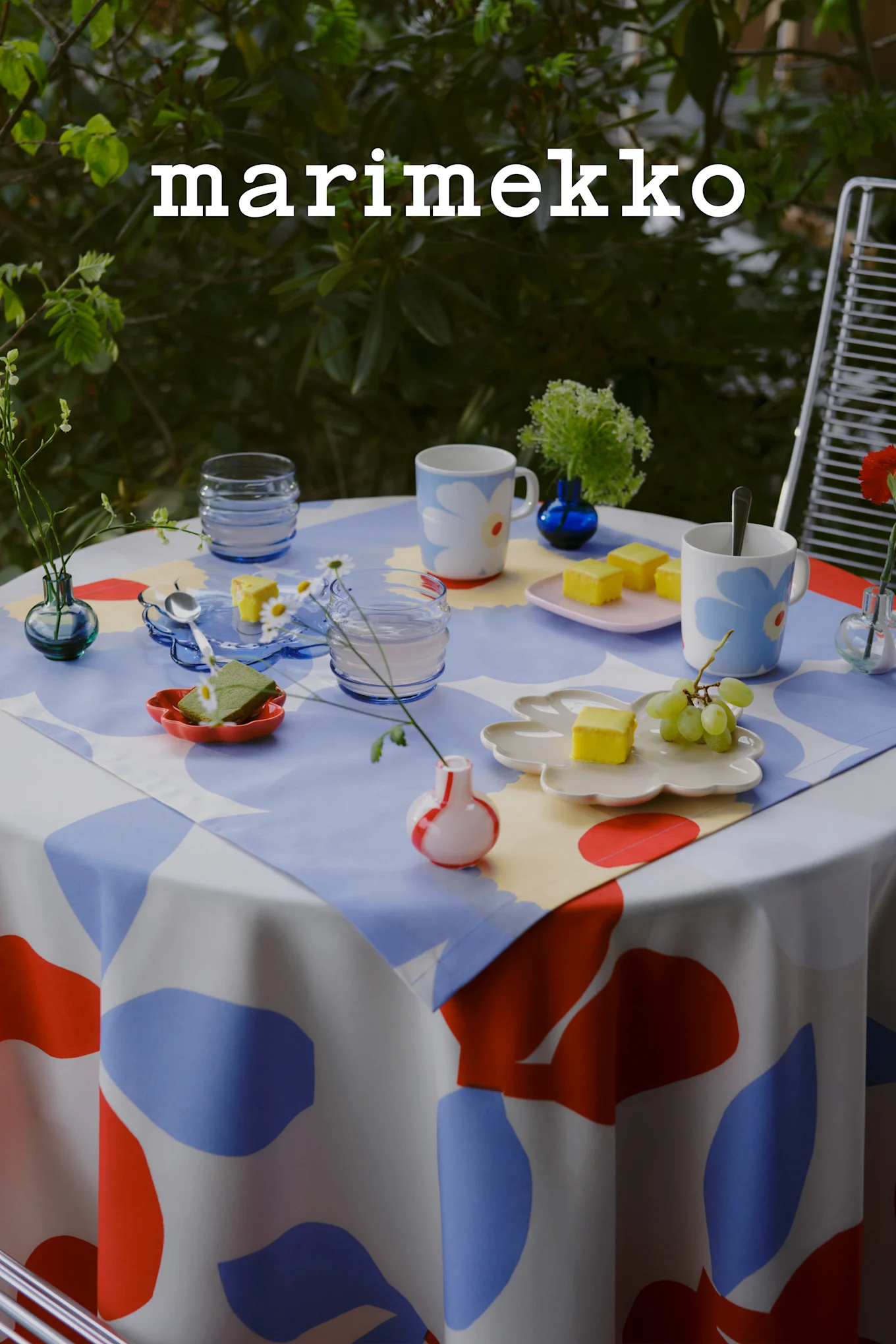 Marimekko logo. Udendørsbord med blå, rød, hvid abstrakt dug, blomstermønstrede krus, kager, druer, små vaser.