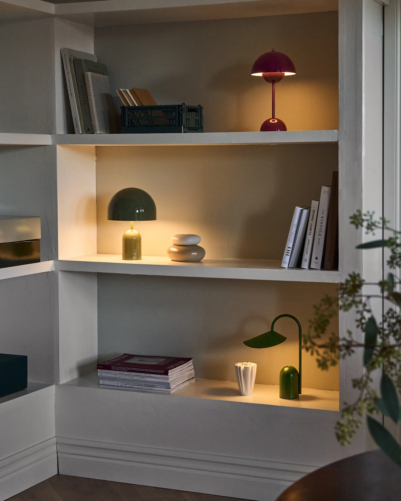 Tre oplyste hylder i en indbygget enhed, der viser røde og grønne svampelamper, en grøn bordlampe, bøger og dekoration.