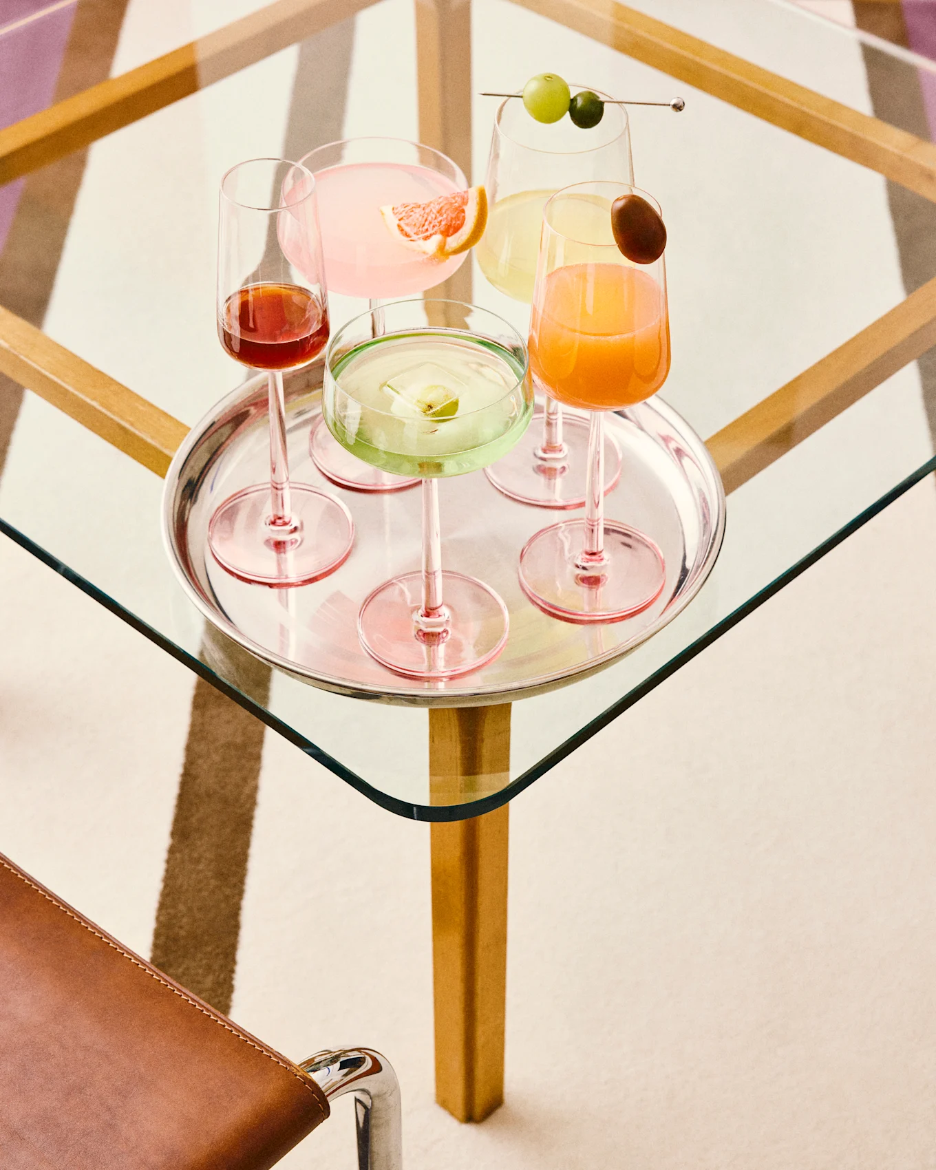 Fem farverige cocktails på en sølvbakke: pink med grapefrugt, grøn med frugtis, gul med druer, orange med pynt og mørkerød, alle på et glasbord.