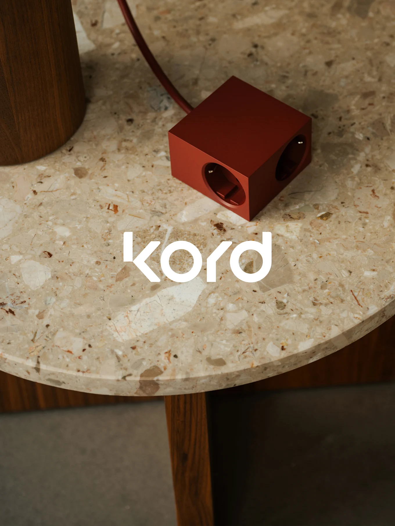 En mørkerød, terningformet stikdåse med to synlige udtag hviler på et rundt marmorbord, med det hvide "kord"-logo.