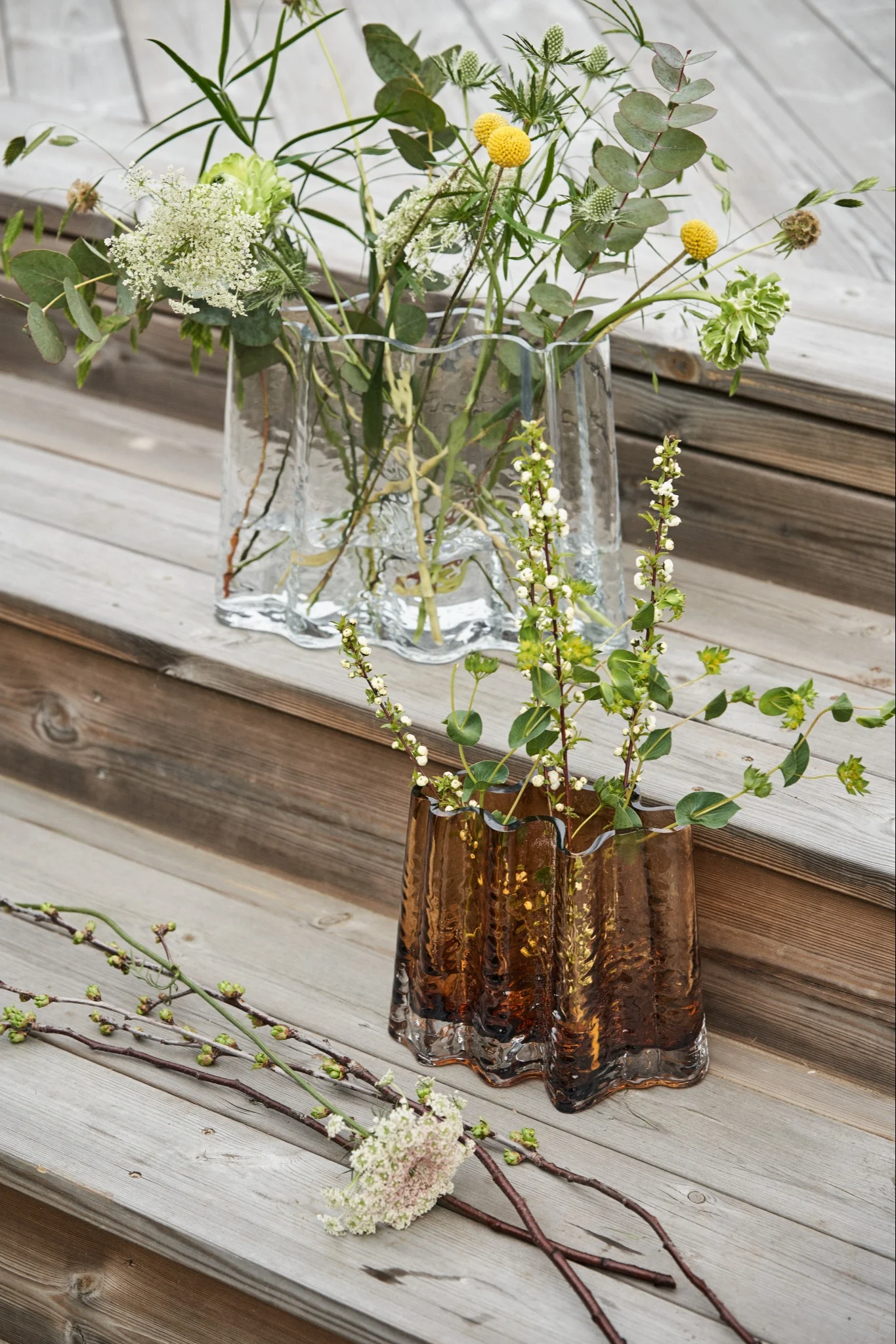 Gry vasen i klar og cognac står på en træ trappe omringet af forårsblomster.