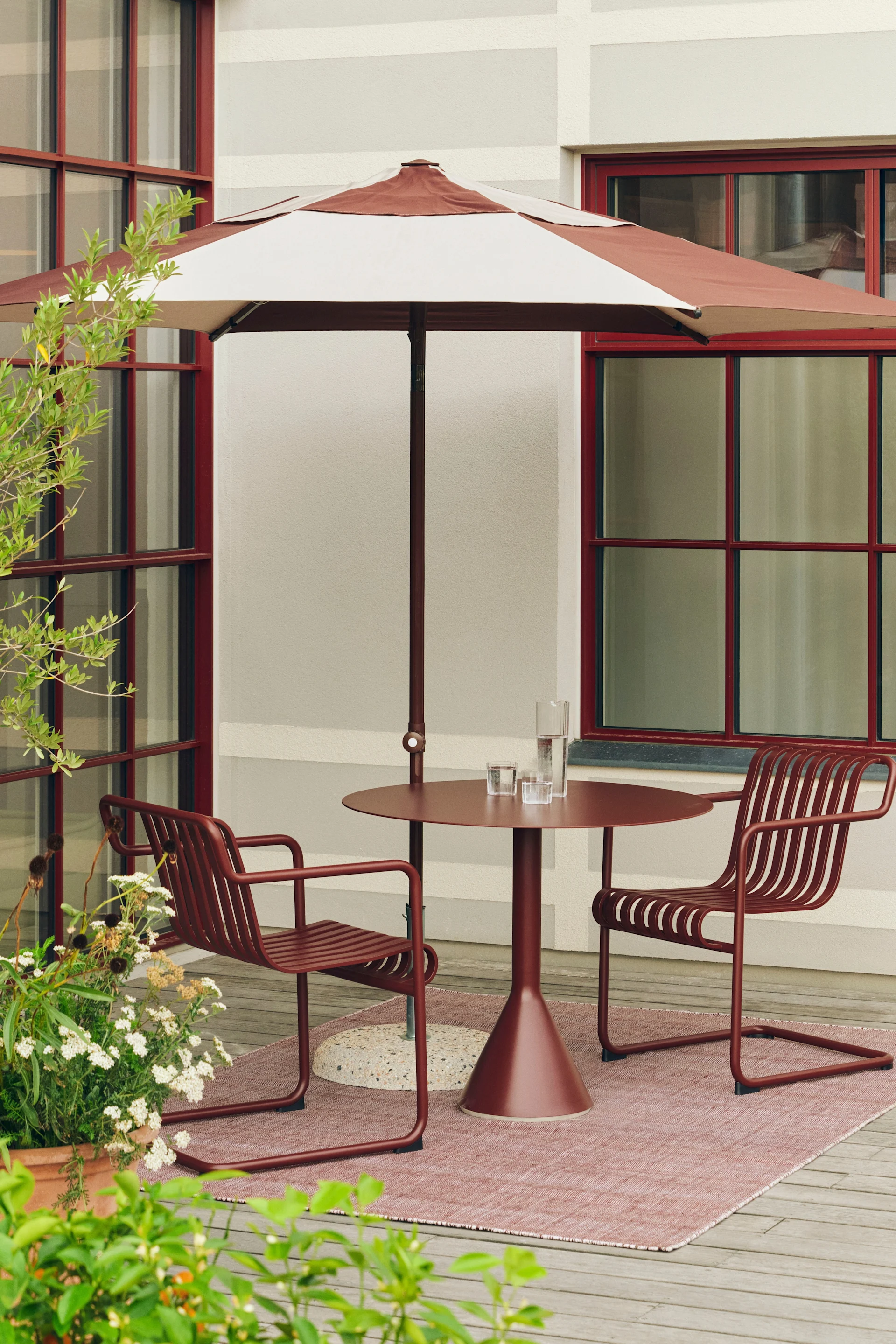Udendørs terrasse med rødbrun stribet parasol, matchende rundt bord og to stole med lameller på et lyserødt tæppe.
