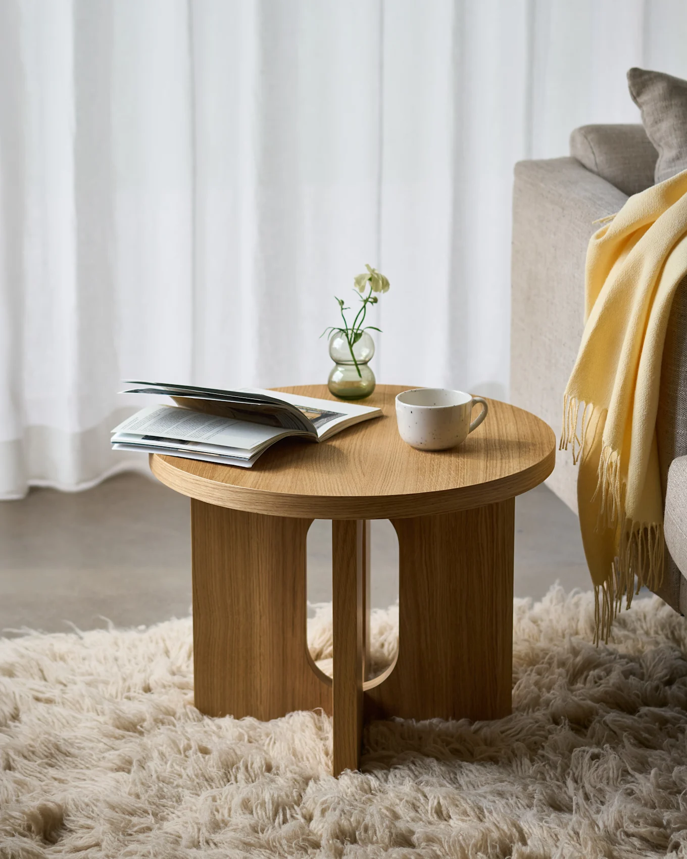 Rundt træsidebord på et fluffy tæppe, med et åbent magasin, en hvid blomst i en vase og en keramikkop, ved siden af en sofa.