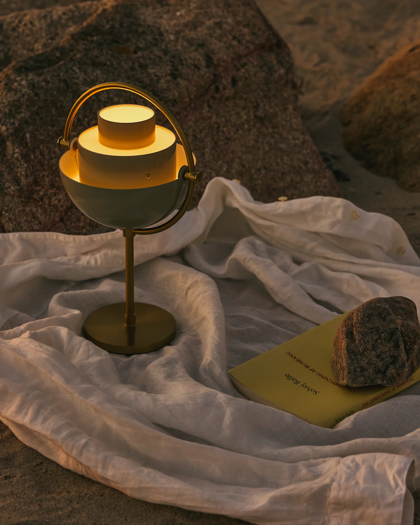 En moderne hvid og messing bordlampe med et glødende lys, en gul bog og en sten på et hvidt klæde på en stenet overflade.