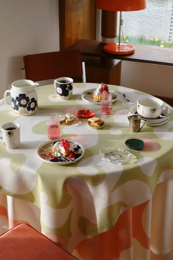 Et rundt bord med en grøn, lyserød og hvid blomstret dug, dækket med sort- og lyserødmønstrede krus, tallerkener, en kande, kager toppet med røde ribs og lyserøde glas.