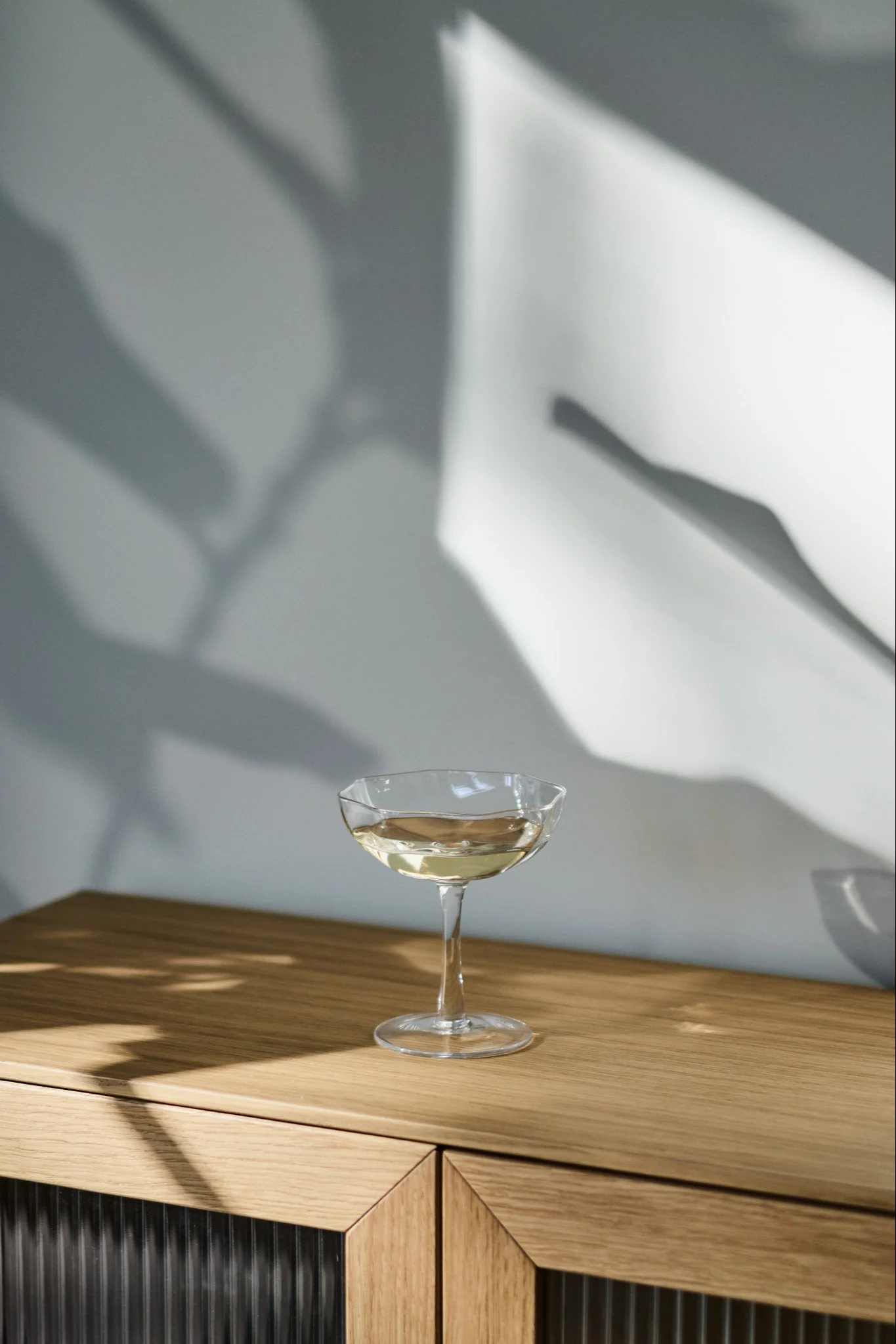Et coupeglas med en lys drink på et træskab, med abstrakte skygger på væggen.