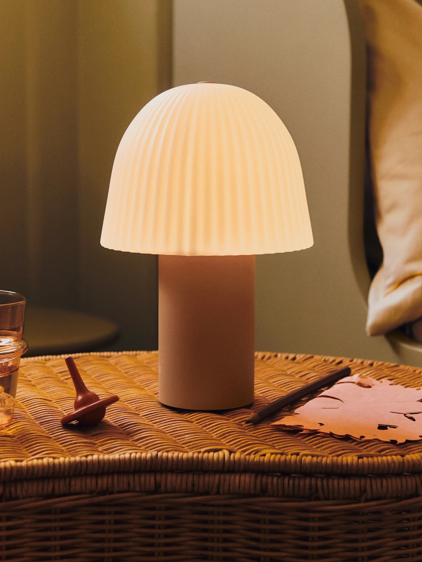En glødende champignonlampe med plisseret skærm på et flet-natbord med et glas vand, en snurretop og papir.