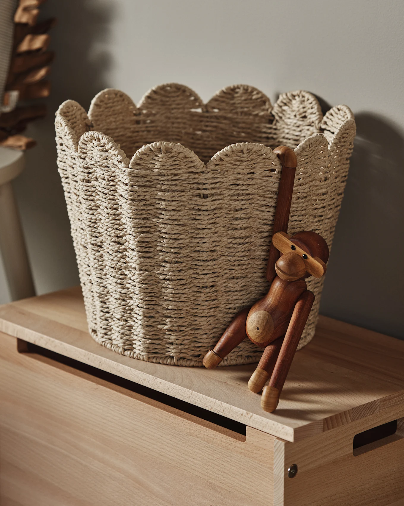 En lys flettet kurv med bølget kant står på en træoverflade, med en træabe-figur hængende fra siden.