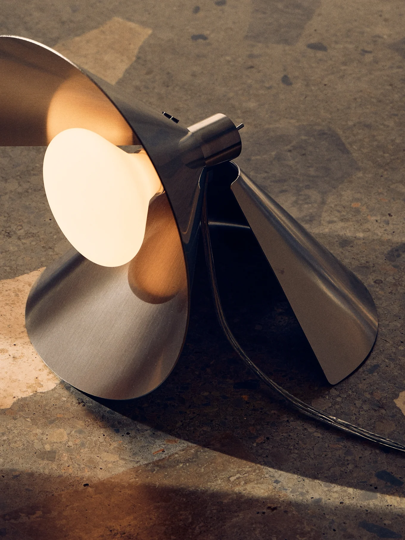 Moderne bordlampe i børstet metal med et skulpturelt, foldet design og en varm, eksponeret pære på en tekstureret overflade.