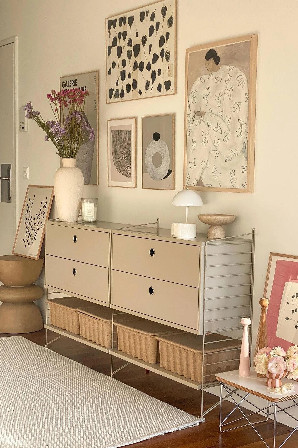 Et moderne værelse med et beige modulopdelt skab, skuffer og opbevaringskurve. En vase med blomster, lampe og abstrakt kunst pryder rummet.