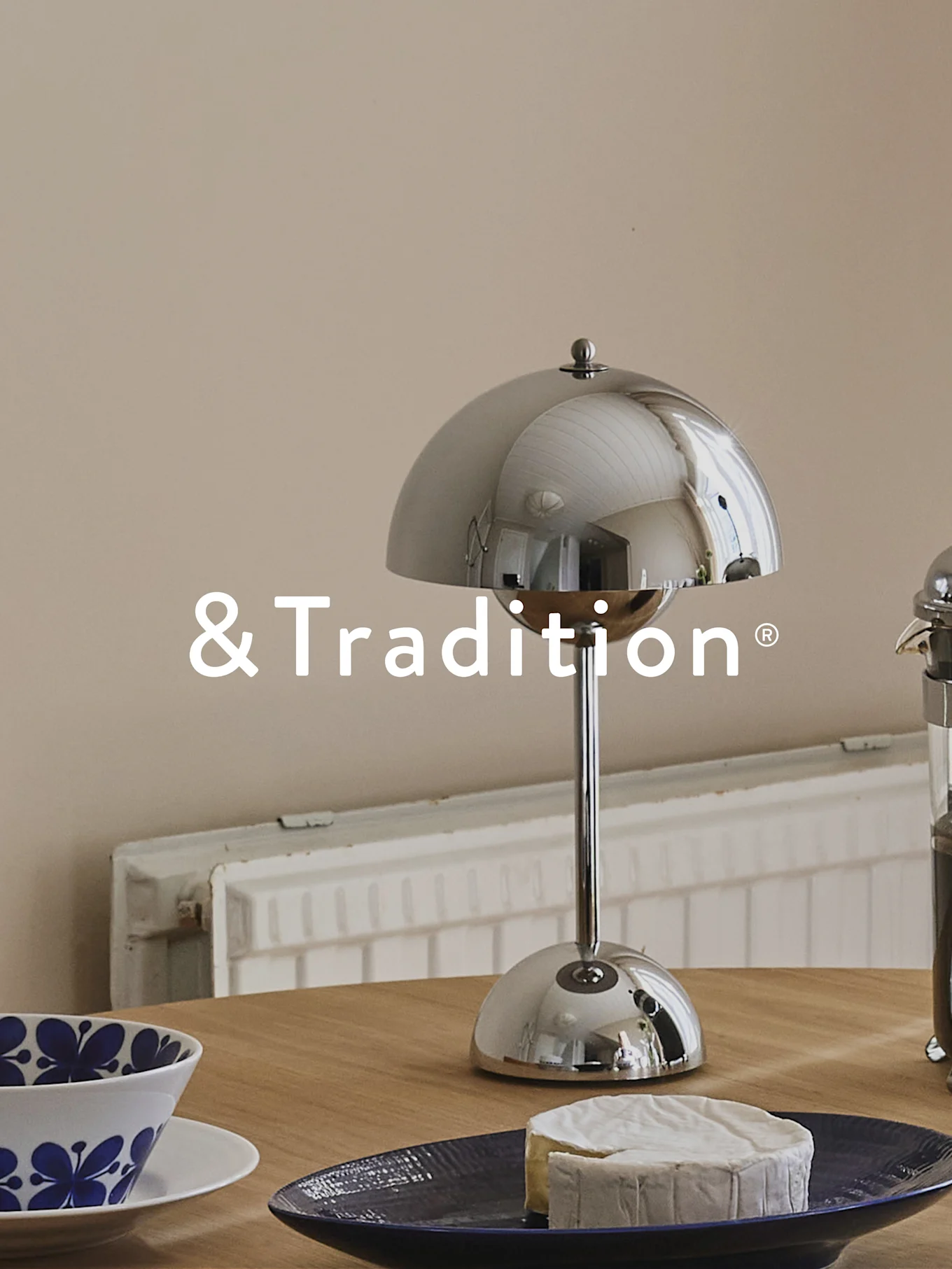 En sølvkuppelformet bordlampe, en rund ost på en mørkeblå tallerken og en blåmønstret skål på et træbord. &Tradition logo.