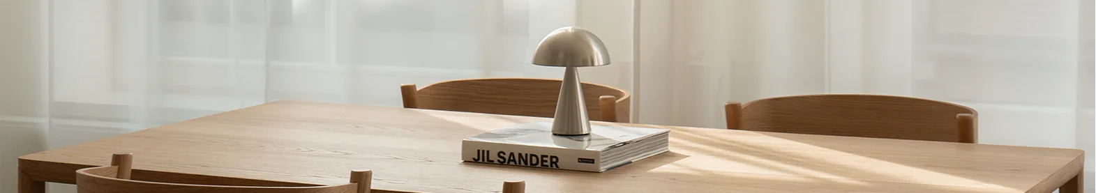 Lyst træ spisebord med fire stole. Sølv svampelampe og bog på bord. Sollys gennem gennemsigtige hvide gardiner.