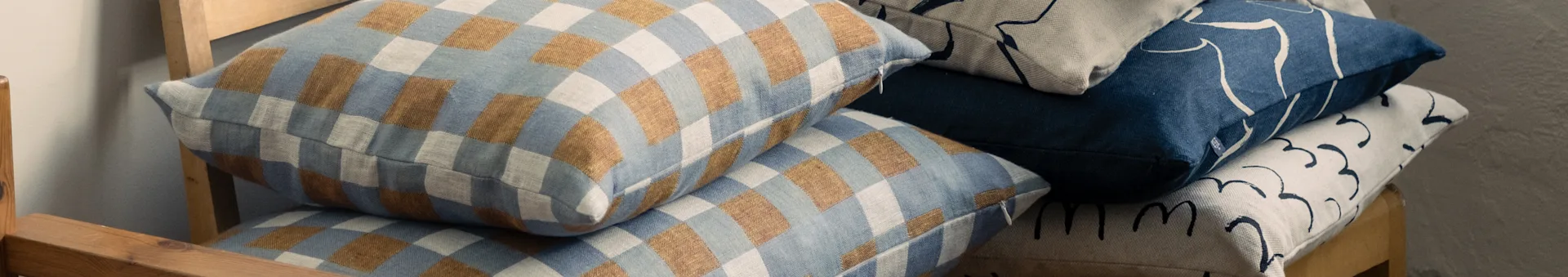 Stakkede puder på en træstol, med ternede blå og brune, samt abstrakte mønstrede blå og beige designs.