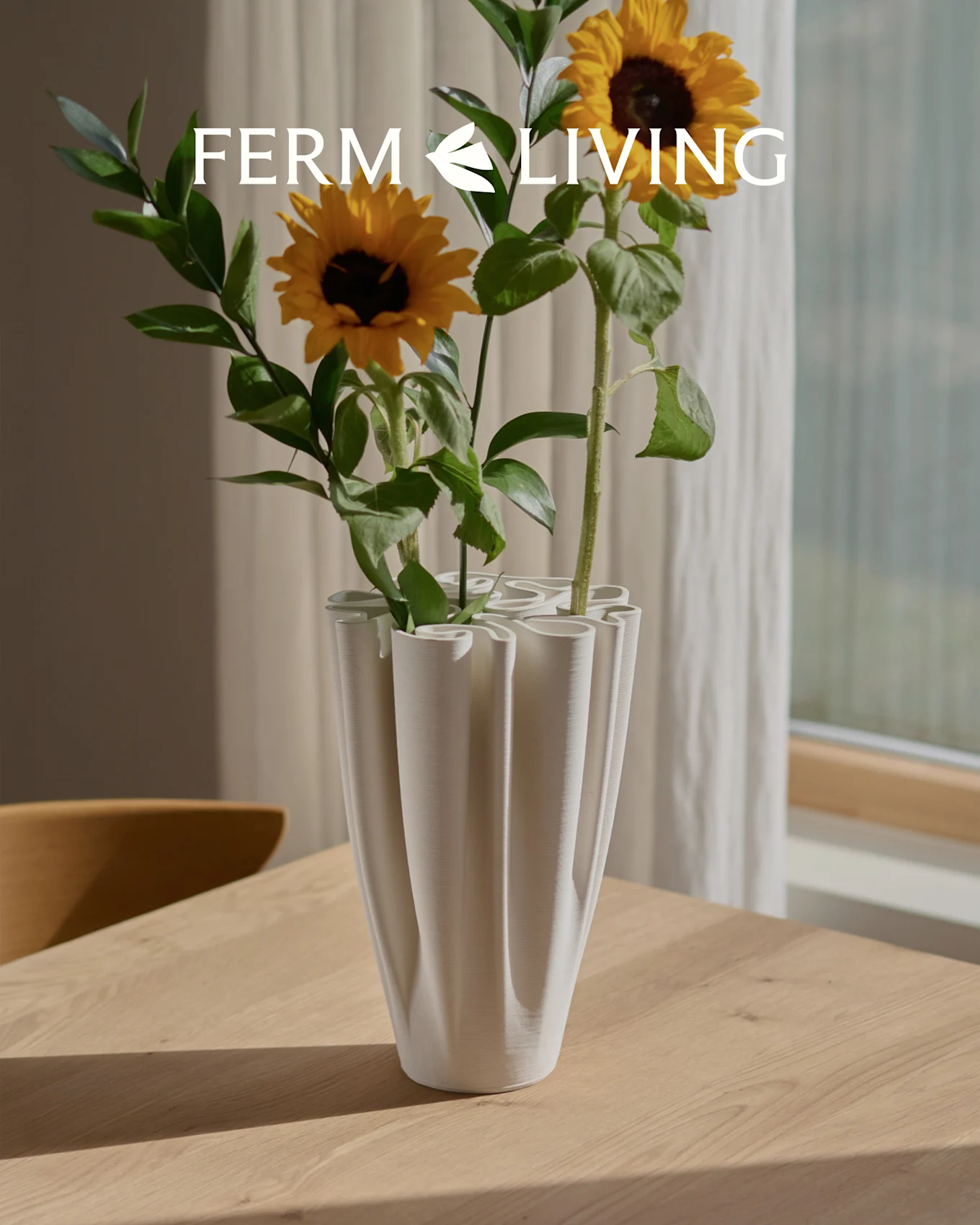Solsikker i en hvid, riflet bølgevase på et træbord. Ferm Living logo overlay.