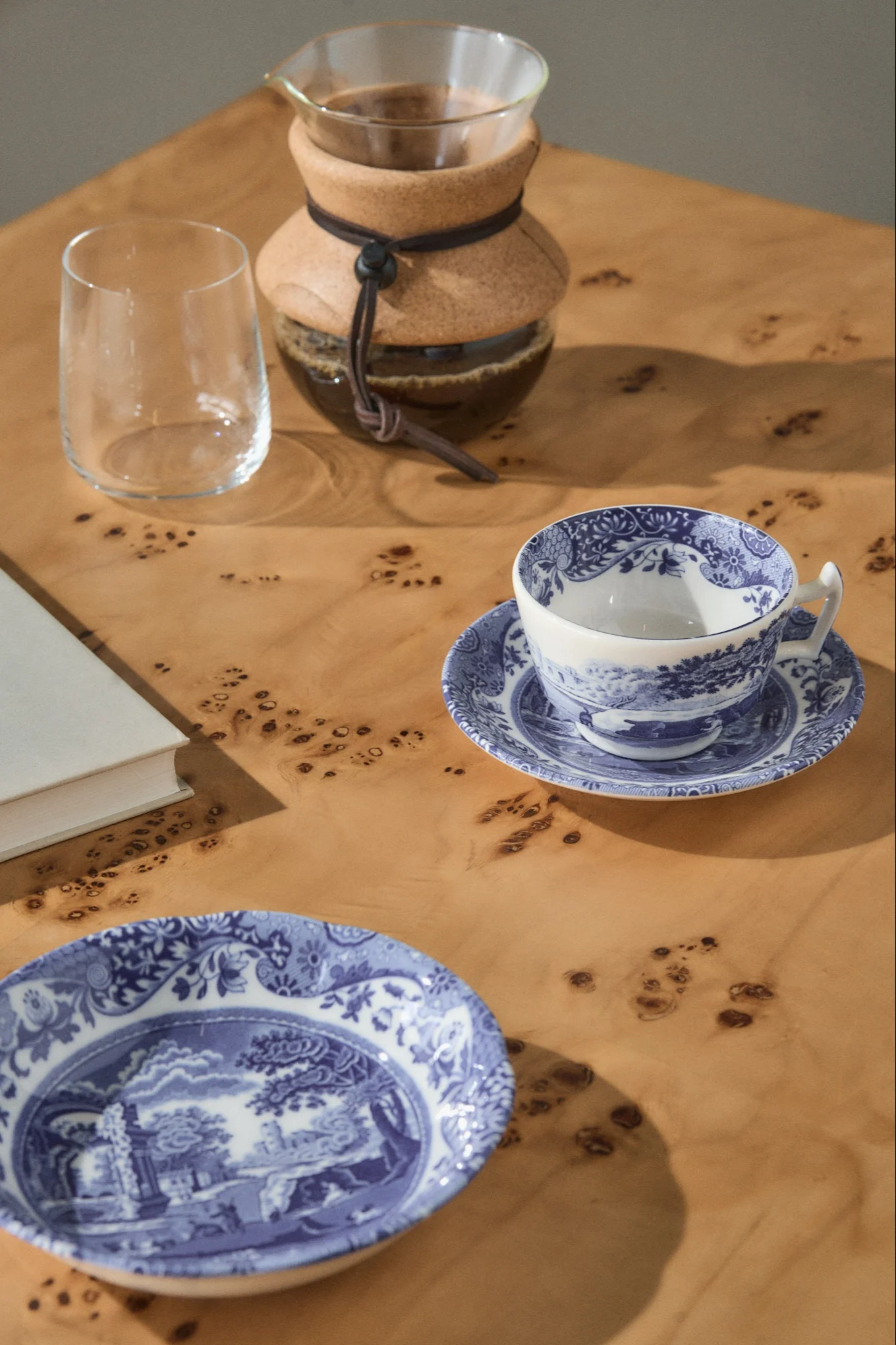 En pour-over kaffemaskine, et tomt glas, en blå- og hvidmønstret tekop på en underkop, og en matchende tallerken på et lyst træbord.
