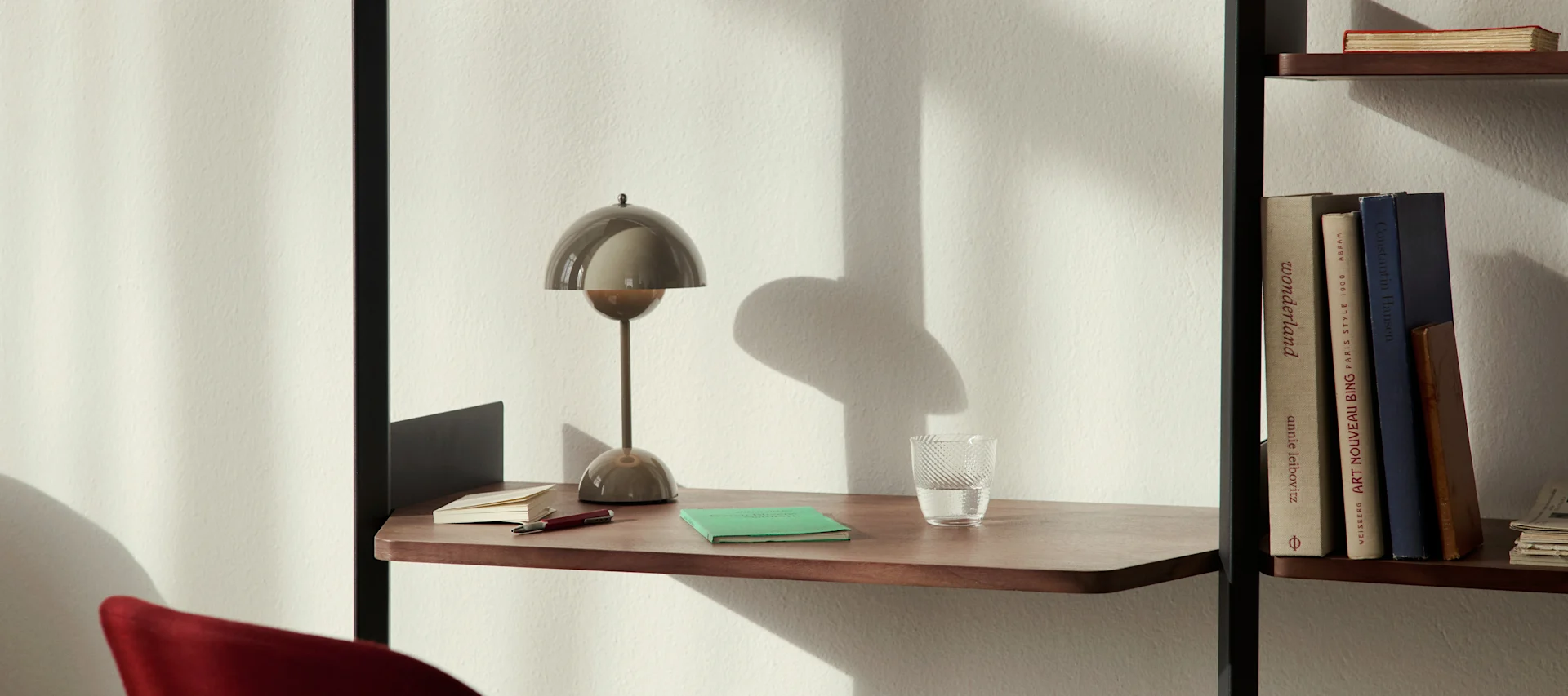 En moderne svampformet skrivebordslampe, et glas vand og notesbøger på en træhylde ved siden af en bogreol.
