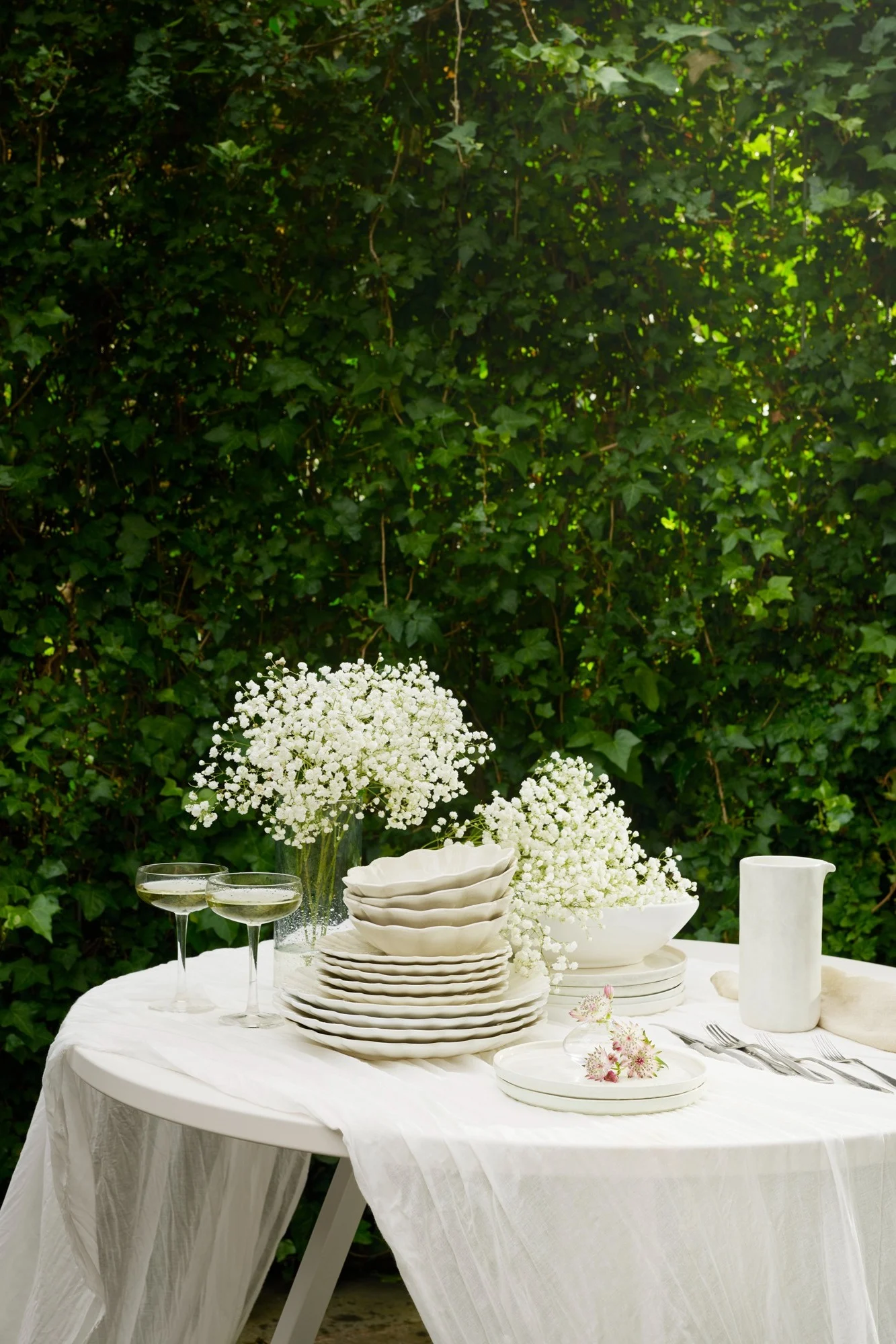 Udendørs bord dækket med hvidt service, brudeslørblomster, champagneglas og bestik på en hvid dug, mod frodige grønne blade.