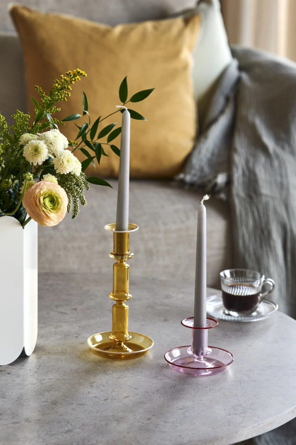 To glas lysestager, en ravfarvet og en lyserød, med grå lys på et bord ved siden af en vase med blomster og en kop kaffe.