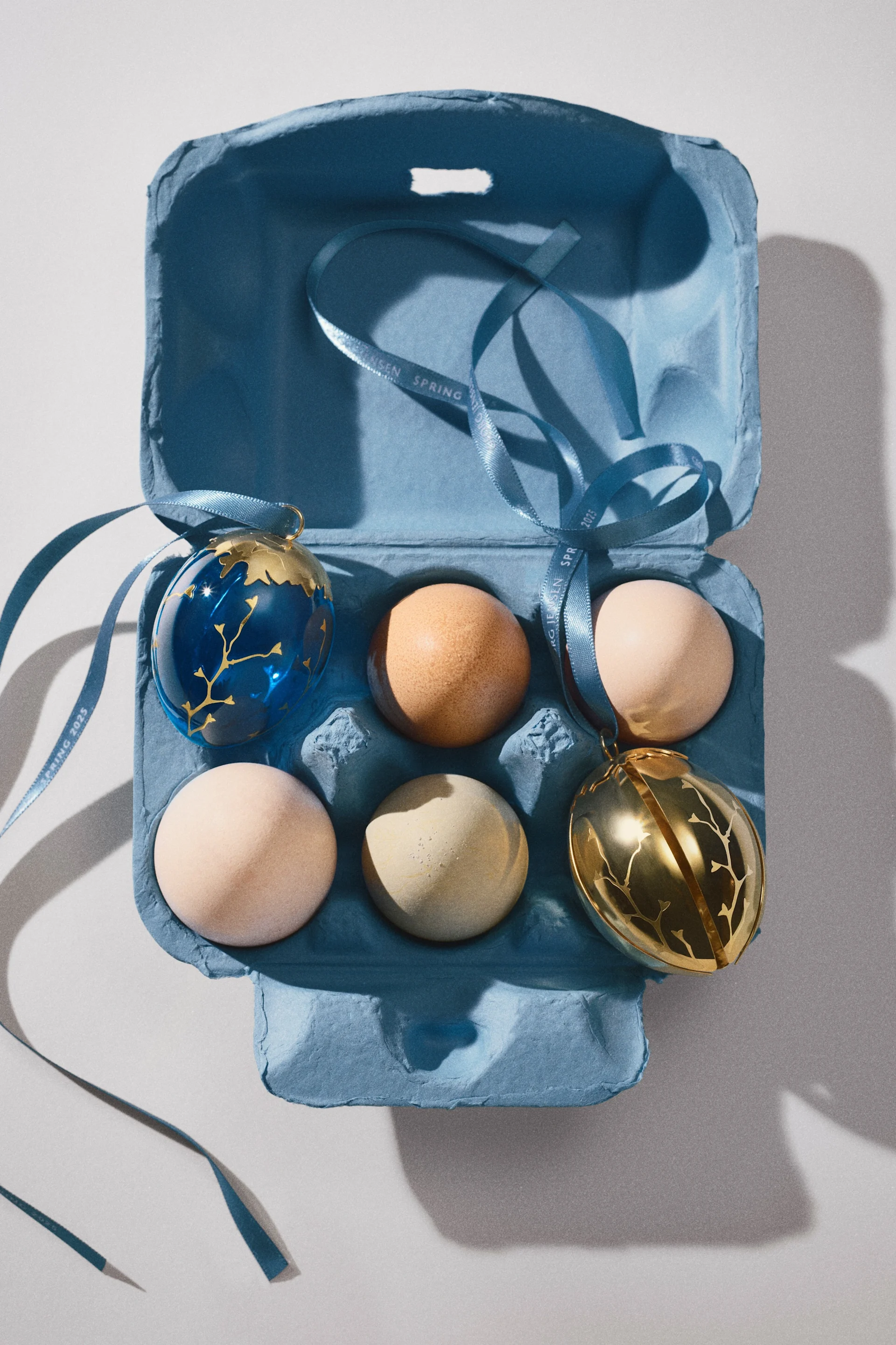 Det gyldne og blå dekorationsæg fra Georg Jensens Spring 2025-kollektion sammen med rigtige æg i en æggeboks.