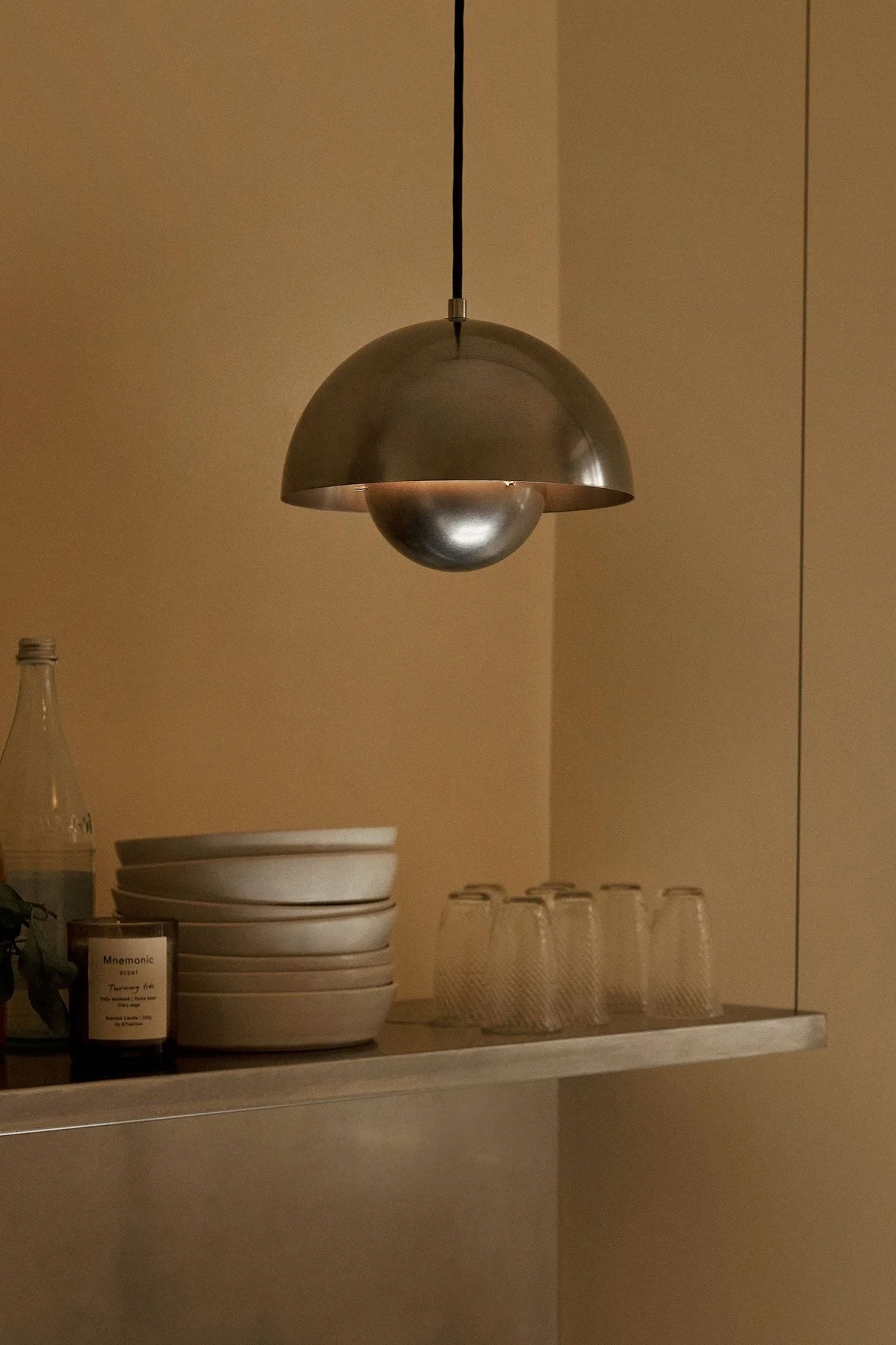 Metallisk kuppelpendellampe, der oplyser en hylde med stablede skåle, klare glas, en flaske og et stearinlys i et varmt rum.