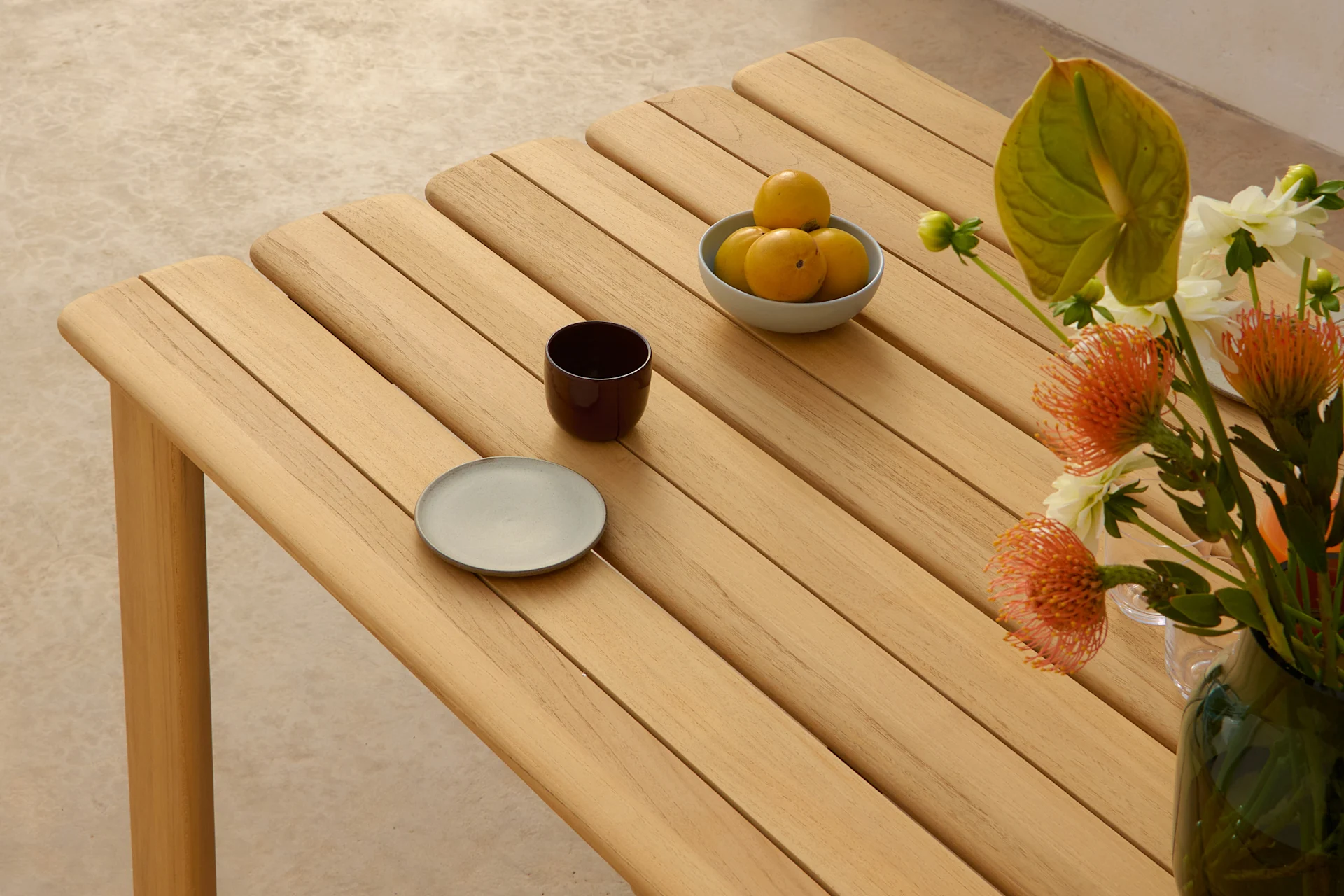 Et lyst trælamelbord med en skål gule frugter, en mørk kop, en grå tallerken og en vase med orange og hvide blomster.