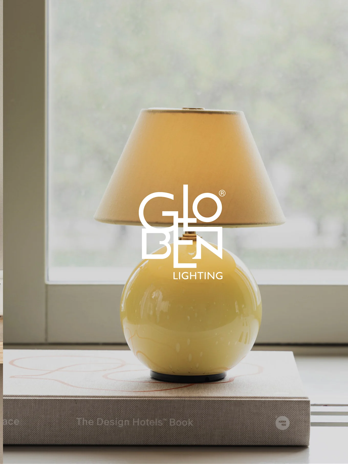 Gul bordlampe med beige skærm, står på bøger ved et vindue.