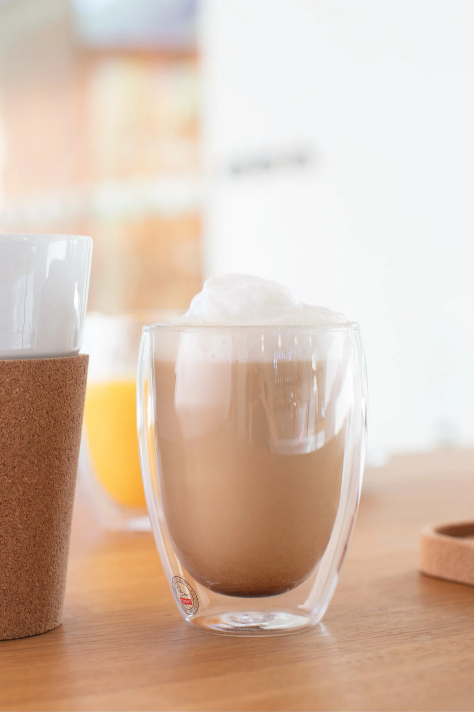 Skummende latte i et dobbeltvægget klart glaskrus på et træbord, med et andet krus med en korkmanchet til venstre og en orange drik i baggrunden.