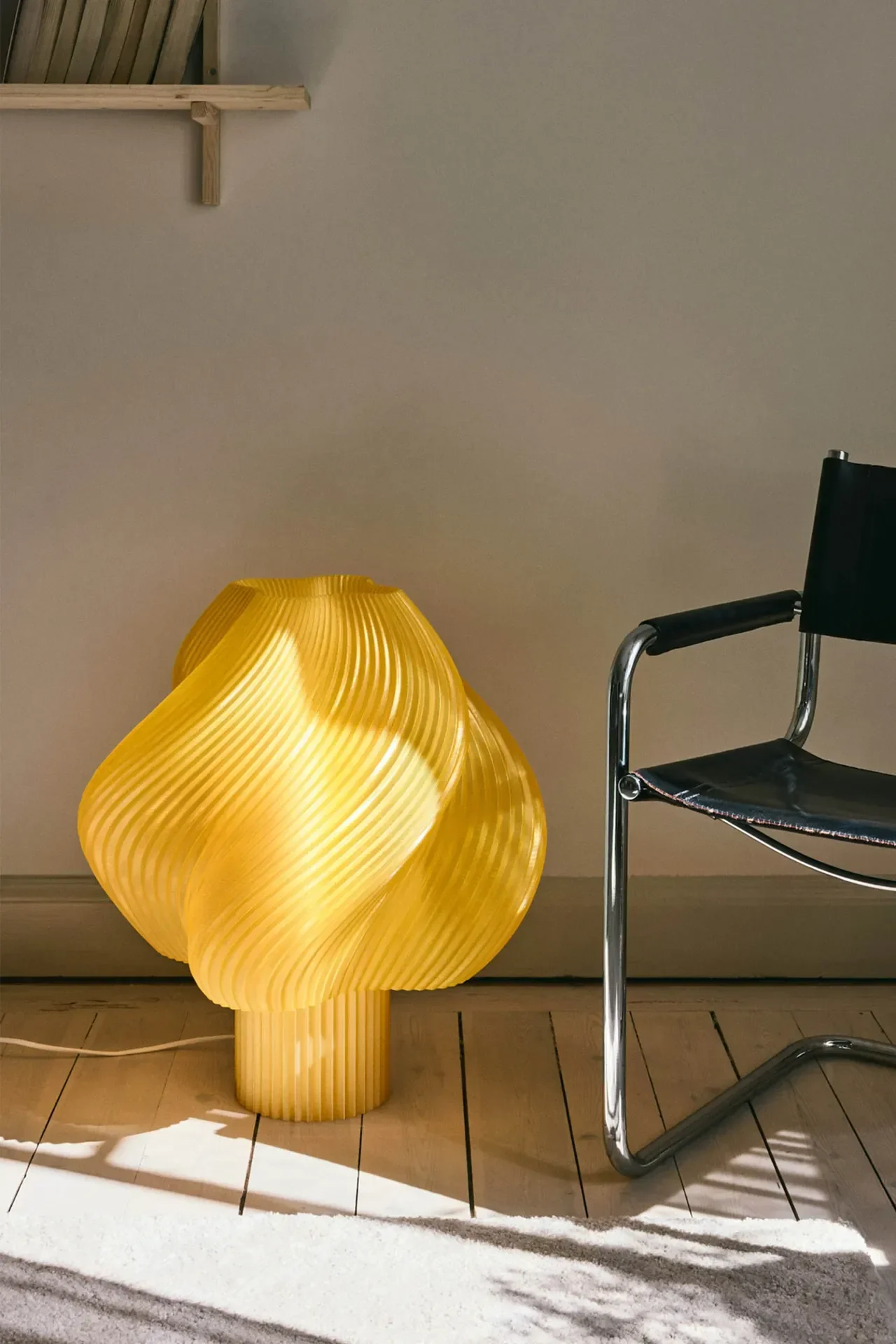 En lys gul, skulpturel bordlampe med et ribbet, bølget design lyser på et trægulv ved siden af en sort læder- og kromstol.