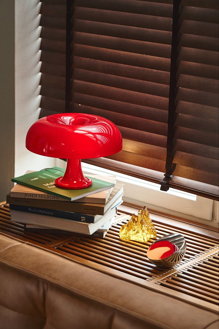 Her ser du en af efterårets fedeste lamper som passer med retro trenden, Nessino bordlampen i postkasserød ovenpå en stak bøger i et retro lignende vindue i mørkt træ.