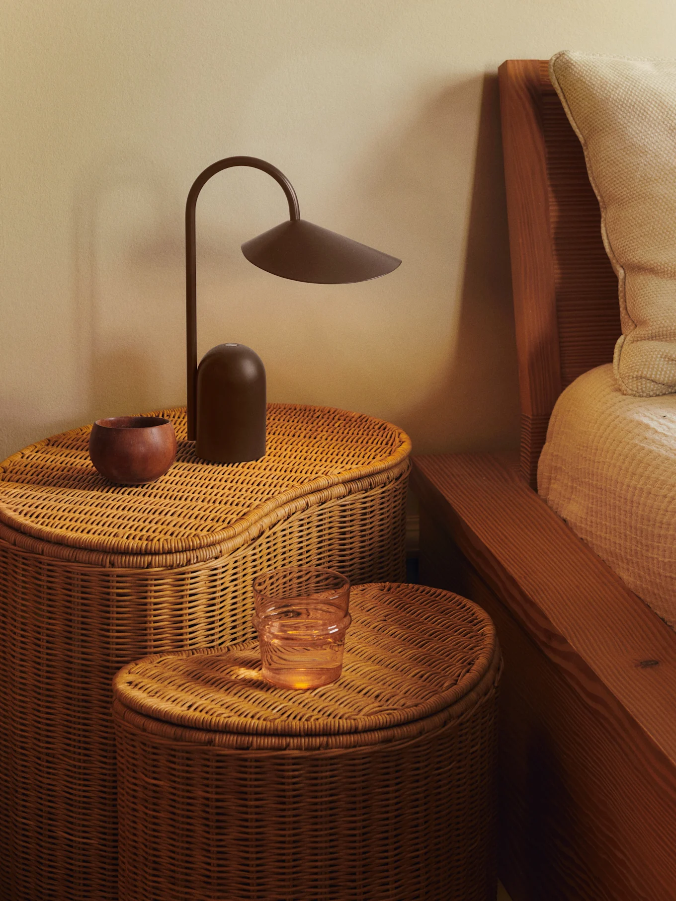 To flettede sengeborde i rattan med en mørkebrun lampe, en træskål og et glas vand.