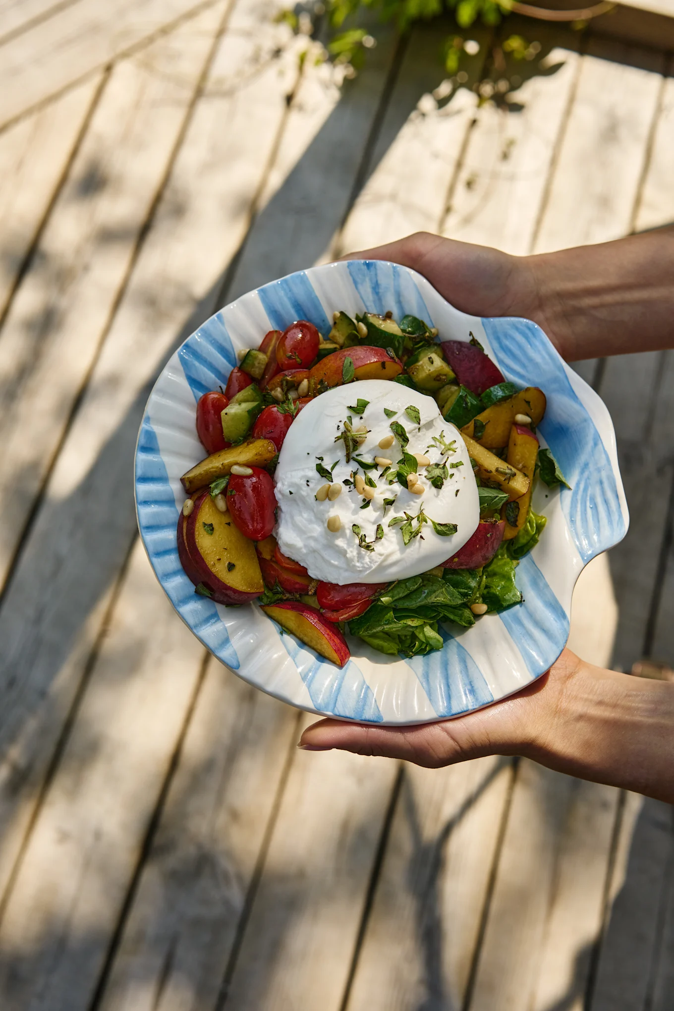 Hænder holder en burrata-salat med tomater, ferskner, agurk og grønt i en stribet skål formet som en musling.