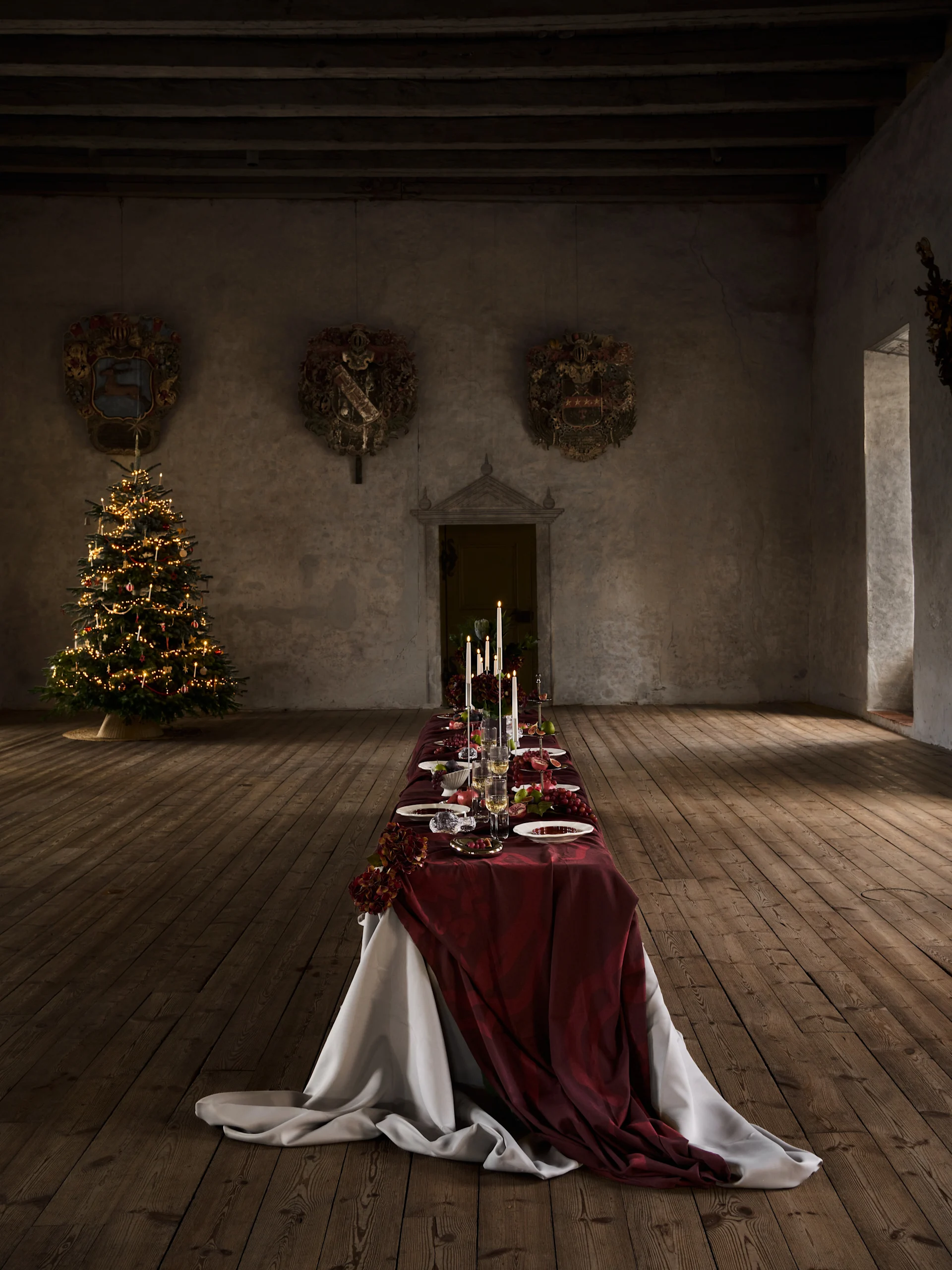 Langt julemiddagsbord med rødt dug, stearinlys, frugt og service, ved siden af et pyntet juletræ i en gammel sal.