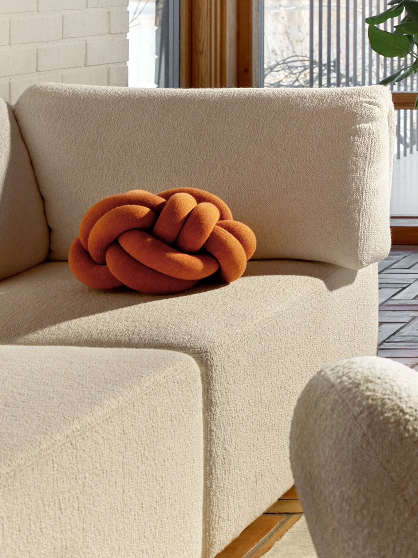 En orange knude-pude på en beige tekstureret sofa med en hvid murstensvæg og et vindue i baggrunden.
